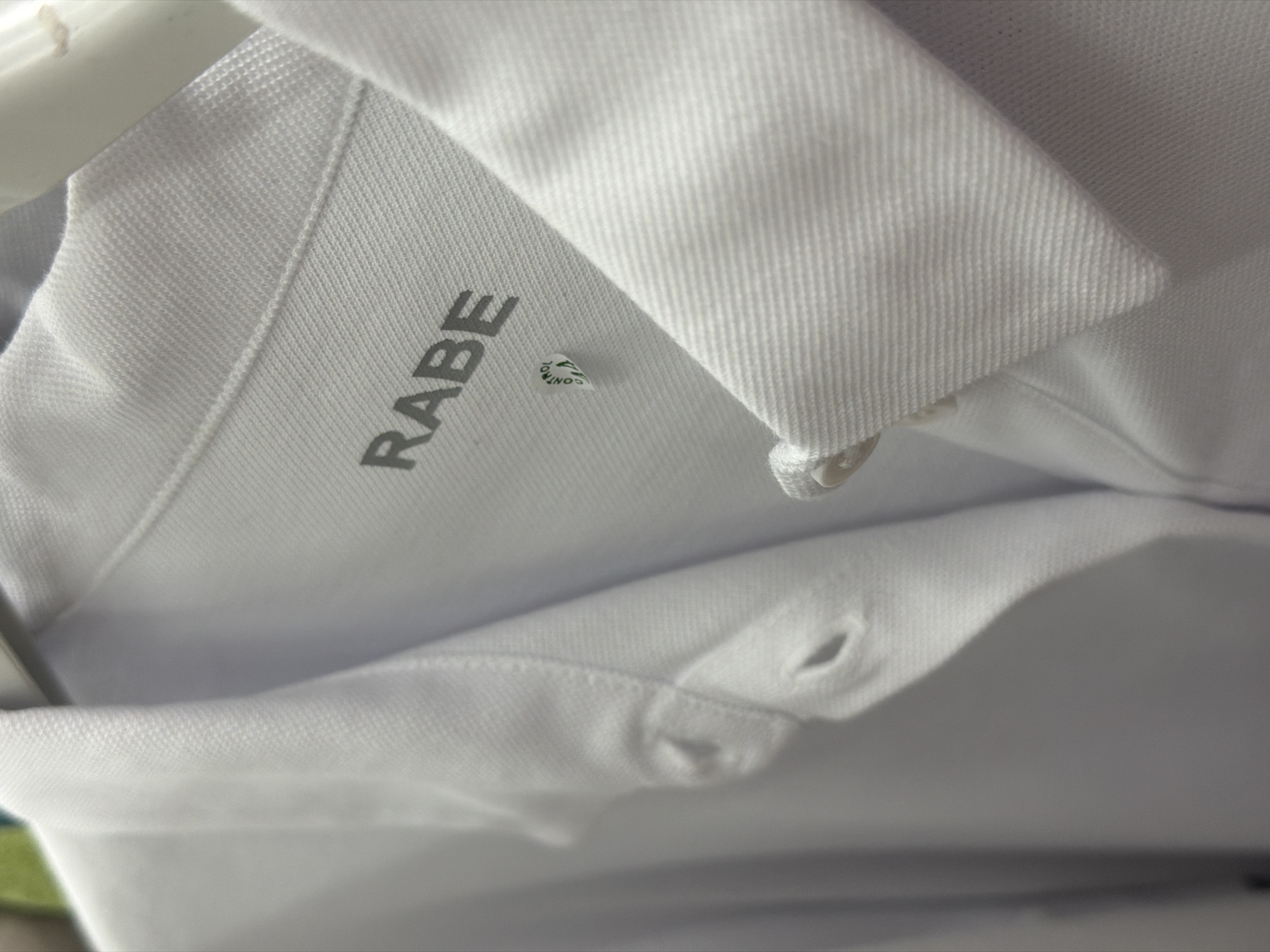 The image shows a close-up of a white shirt with a black logo that reads "RABE" on the collar. There is also a small green and white sticker on the shirt. The shirt appears to be hanging on a clothes hanger. The image is in focus and well-lit. There are no people or any indication of a specific location in Svilengrad, Bulgaria. The scene is a static depiction of clothing.