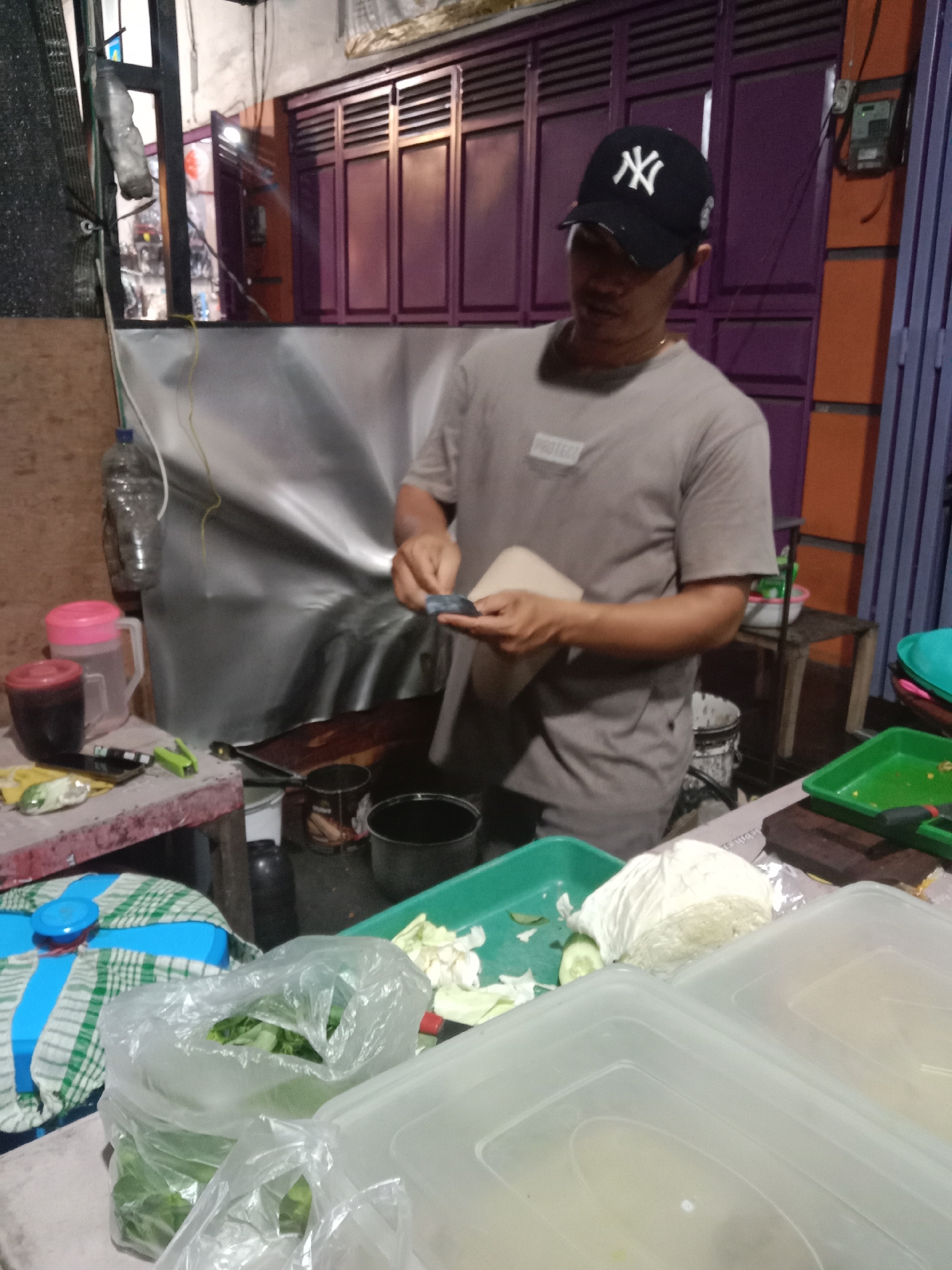 The image depicts a male individual preparing food at a street stall or small establishment in Cangkring, Indonesia. The man, wearing a black baseball cap with a white "NY" logo and a light brown t-shirt with a "PROTEC" patch, is positioned centrally, facing downwards. He is using a small, dark implement in his right hand to work on a light-colored, rectangular food item or wrapper held in his left hand. A thin gold-colored chain is visible around his neck.
To his immediate left, a rustic wooden table holds various items: a red lidded container, a clear plastic pitcher with a pink lid, a green disposable lighter, a shiny metallic object, and a dark, possibly leather, object. A clear plastic bottle hangs from a wire above this table. Behind the table, a silvery-grey tarp or sheet forms a backdrop. Further left, a window shows blurred reflections.
In the mid-ground, a dark frying pan rests on what appears to be a small stove or burner. Several dark pots and a white bucket are visible beneath the work surface.
The foreground on the left features a blue and white checkered cloth covering a round blue container. Beside it are multiple clear plastic bags filled with green leafy vegetables. To the right, white rectangular plastic containers with lids are present. A green plastic tray contains chopped white cabbage and cucumber slices, alongside a dark cutting board and a dark-handled knife. A small, light-colored item wrapped in foil is also on the surface.
The background consists of purple corrugated metal shutters or doors, likely belonging to a building. To the right of these shutters, an orange wall segment is visible, with a grey electrical meter box mounted on it. Various wires are strung across the scene. The overall lighting suggests artificial illumination in an evening or nighttime setting.