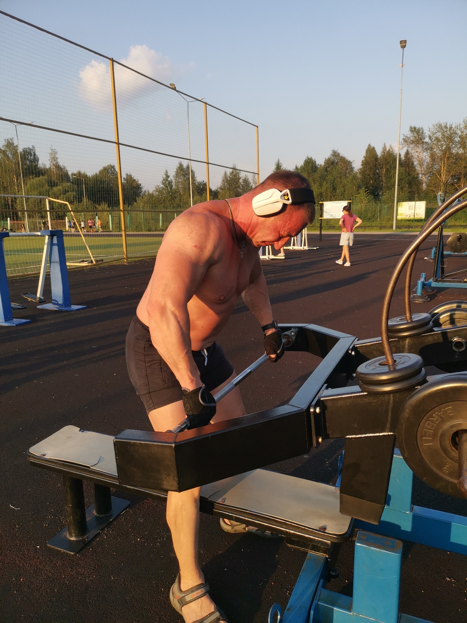 This image depicts an outdoor fitness area, likely a sports ground, in the city of Kizel, Russia. The primary subject is a shirtless, muscular man with a tan, wearing black shorts, black gloves, and white headphones. He is engaged in an exercise on a piece of weightlifting equipment, gripping a metal bar. The equipment is primarily black and blue. In the background, several other individuals are visible, including a person in a pink shirt and grey shorts standing near a sign. A sports net, a lamppost, trees, and a clear blue sky with a few clouds complete the environment. The lighting suggests it is late afternoon or early evening.