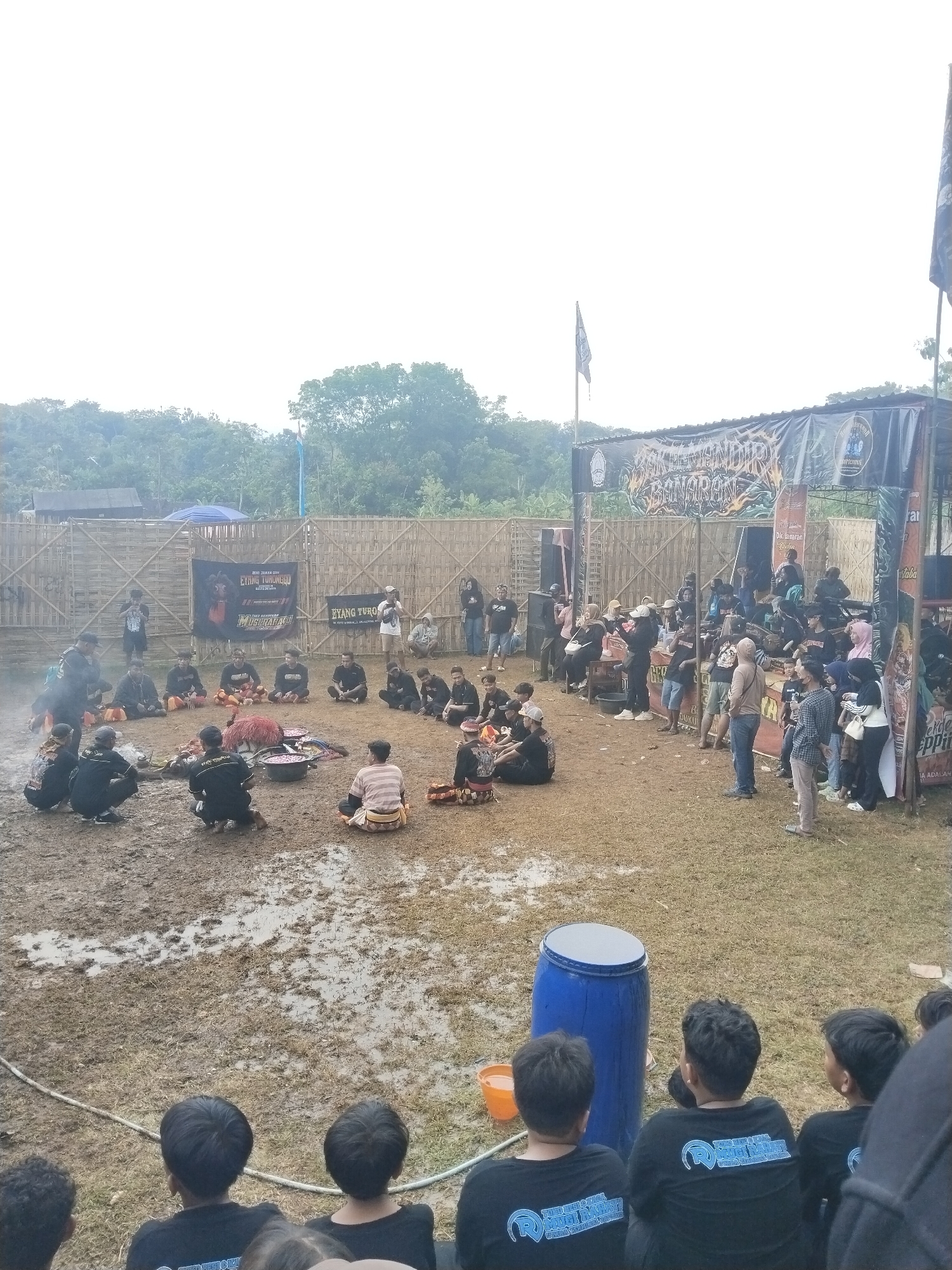 The image captures an outdoor scene at Gentungan, Indonesia, featuring a muddy ground where a group of people sit in a circle. At the center of the circle, there appears to be some type of performance or ritual taking place. Smoke emanates from the center. A small crowd of onlookers surrounds the central area, including people standing and seated. A banner and stage are set up behind the crowd, featuring the text "EYANG TURUNGGU EYANG TURO" and "MUGI RARACU MUGI RAHAT?" An Indonesian flag is present. At the bottom of the image, some people with t-shirts are seated, facing the performance. A blue barrel and an orange bucket are nearby. The surrounding environment includes trees and what appears to be a fence.