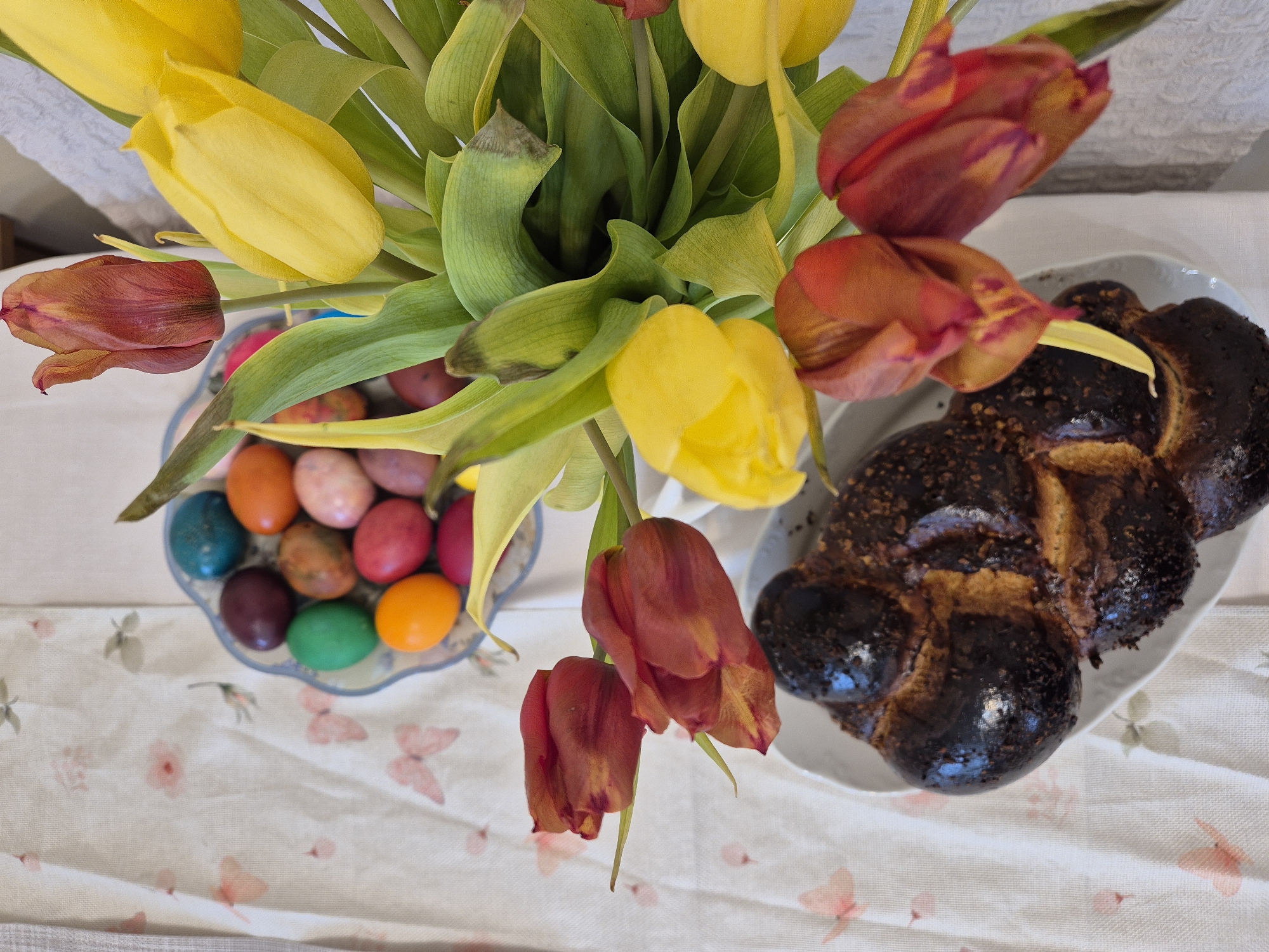 The image is a top-down shot of a table setting with an Easter theme. A bouquet of yellow and red tulips sits centrally in the frame. To the left of the flowers is a shallow, scalloped glass bowl filled with dyed Easter eggs in various colors including blue, orange, pink, red, purple, brown, and green. To the right of the flowers is an oval serving dish with a braided loaf of bread that appears to be somewhat burnt. The table is covered with a patterned tablecloth featuring butterfly and floral motifs. The location is not discernable from the image.