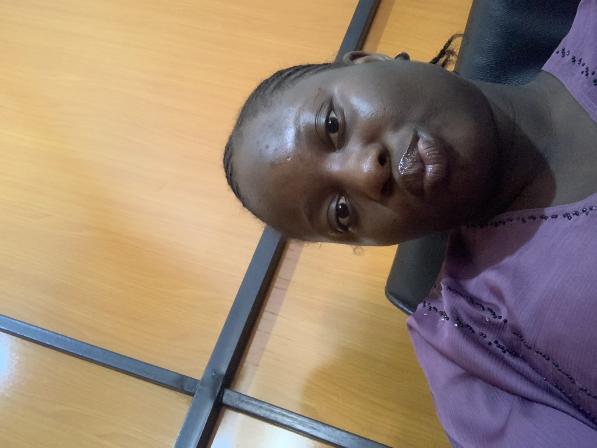 A close-up, waist-up, indoor selfie shows a Black woman with braided hair and a purple blouse with beaded embellishments. The woman is looking at the camera with her lips puckered. She is seated in what appears to be an office or conference room, as indicated by the wood-paneled walls and dark chair. The lighting is warm, suggesting it may be artificial. The woman's face is the focal point of the image, with her eyes open and looking directly at the viewer. Her skin has a dewy sheen, and her lips are glossy. The background is softly blurred, further emphasizing the subject.
