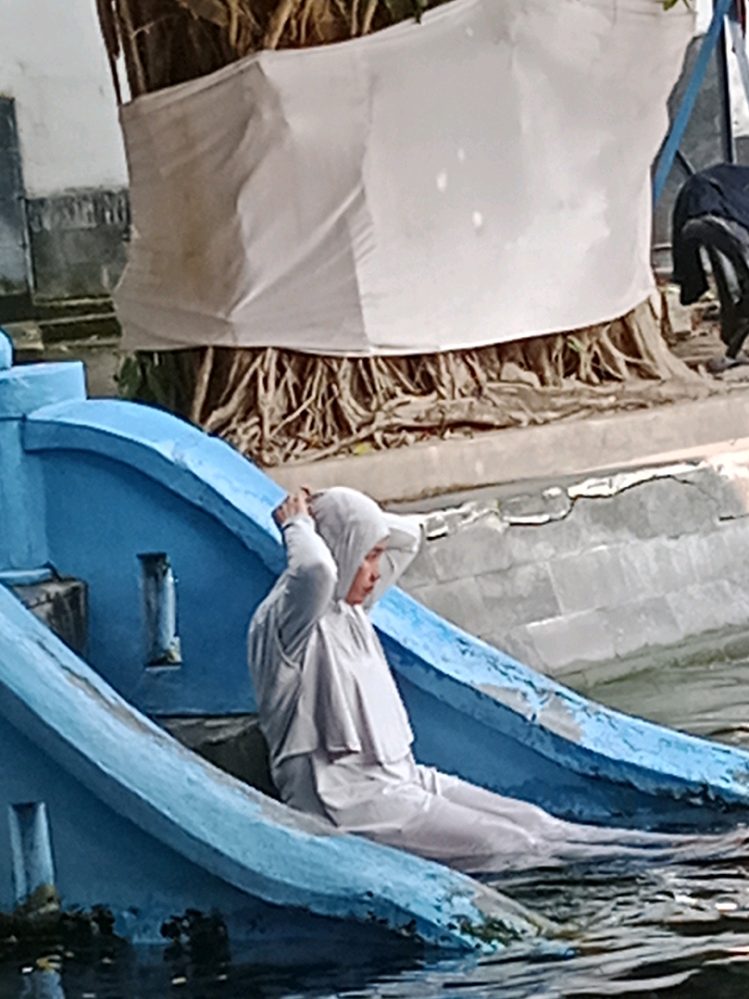 In Jentekan, Indonesia, a person wearing a white hijab and a white garment is seated in a blue structure that appears to be submerged in water. The person is adjusting their hijab. The blue structure has an arched opening. In the background, a tree trunk is wrapped in a white cloth, and there are some loose strands of material at the base of the tree. The water is murky, and reflections are visible on its surface.