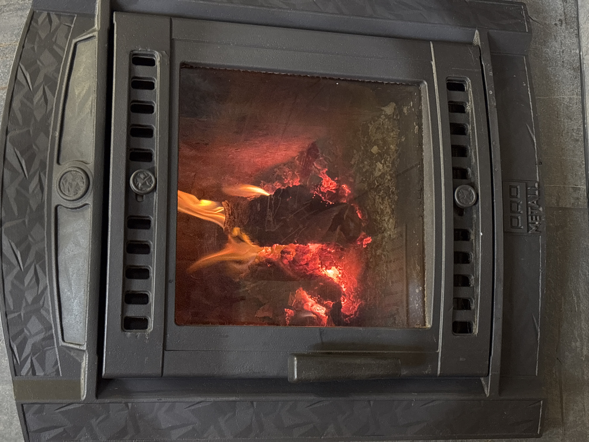 The image shows a close-up, frontal view of a black, cast-iron fireplace with a visible fire burning inside. The fireplace has a decorative, textured pattern on its upper sections and a glass door through which the flames and glowing embers are seen. A handle is visible on the left side of the fireplace door. Below the glass, there are ventilation slots. At the bottom of the fireplace, there is a nameplate with the inscription "PRO METALL". The fireplace is set against a grey tiled wall. The scene is indoors, likely in a residential setting, and the presence of the fireplace suggests a cool climate, consistent with Chaikovsky, Russia. There are no people or animals present in the image.