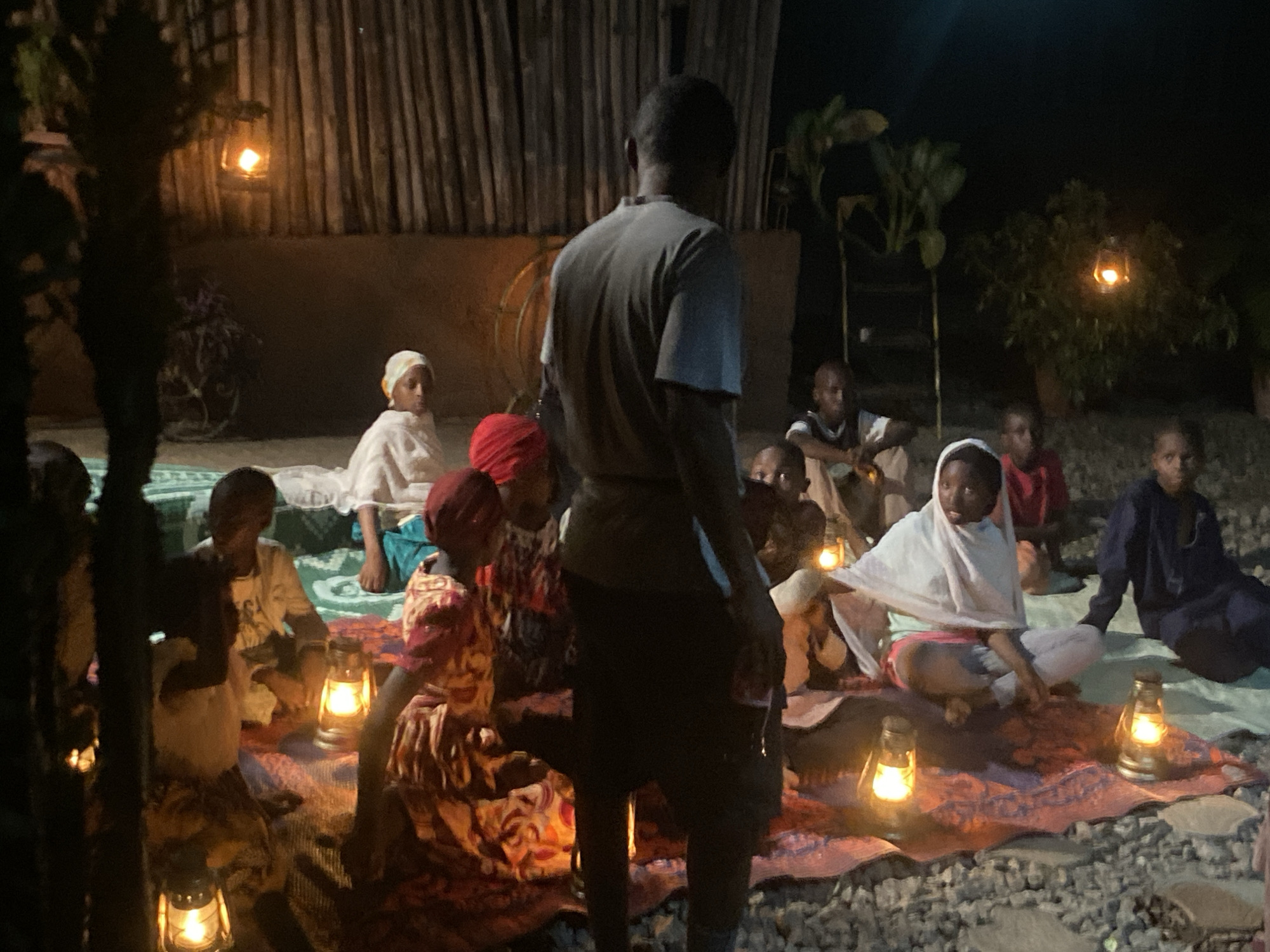 A group of people, including children, sit in a semi-circle on a woven mat outdoors at night, illuminated by several oil lamps on the ground and hanging in trees and on a wall. One adult man stands in the foreground with his back to the camera. The ground appears to be covered in small rocks. The location is Barden Yana, Nigeria.
