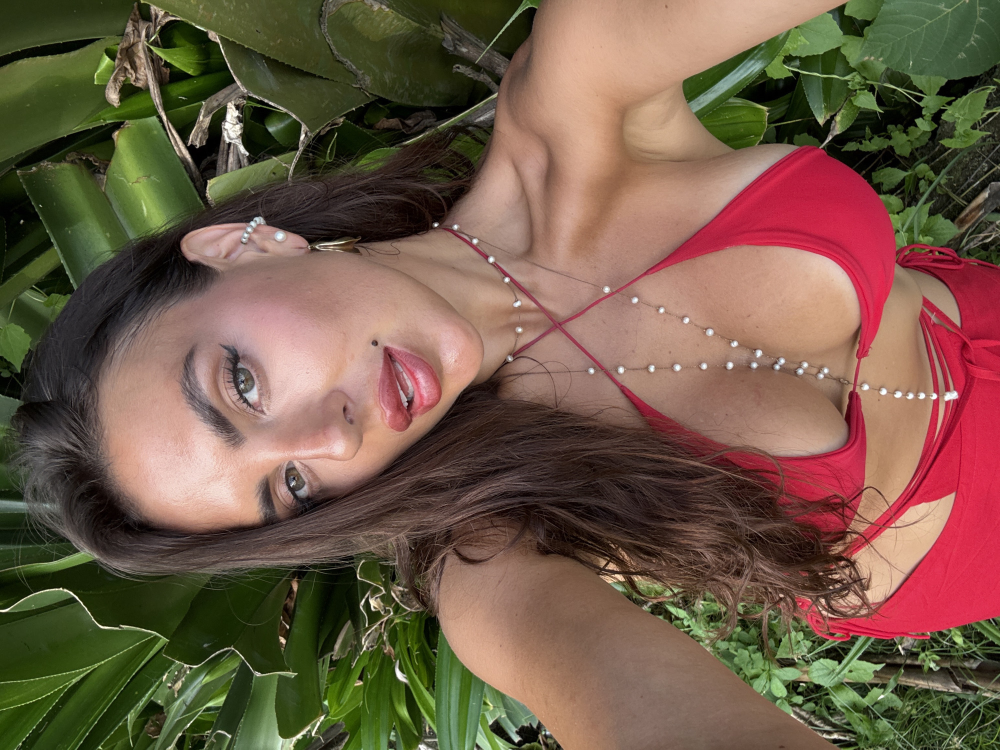 A young woman with long, brown hair and fair skin lies among green, broad-leafed plants, possibly agave. She wears a red bikini top adorned with a delicate pearl-like body chain. The location is likely in Mengwi, Indonesia, suggested by the type of foliage. Her makeup includes lip gloss and eyeliner. She has a mole on her left cheek. Her ear has a pearl cuff earring and a gold pendant earring.