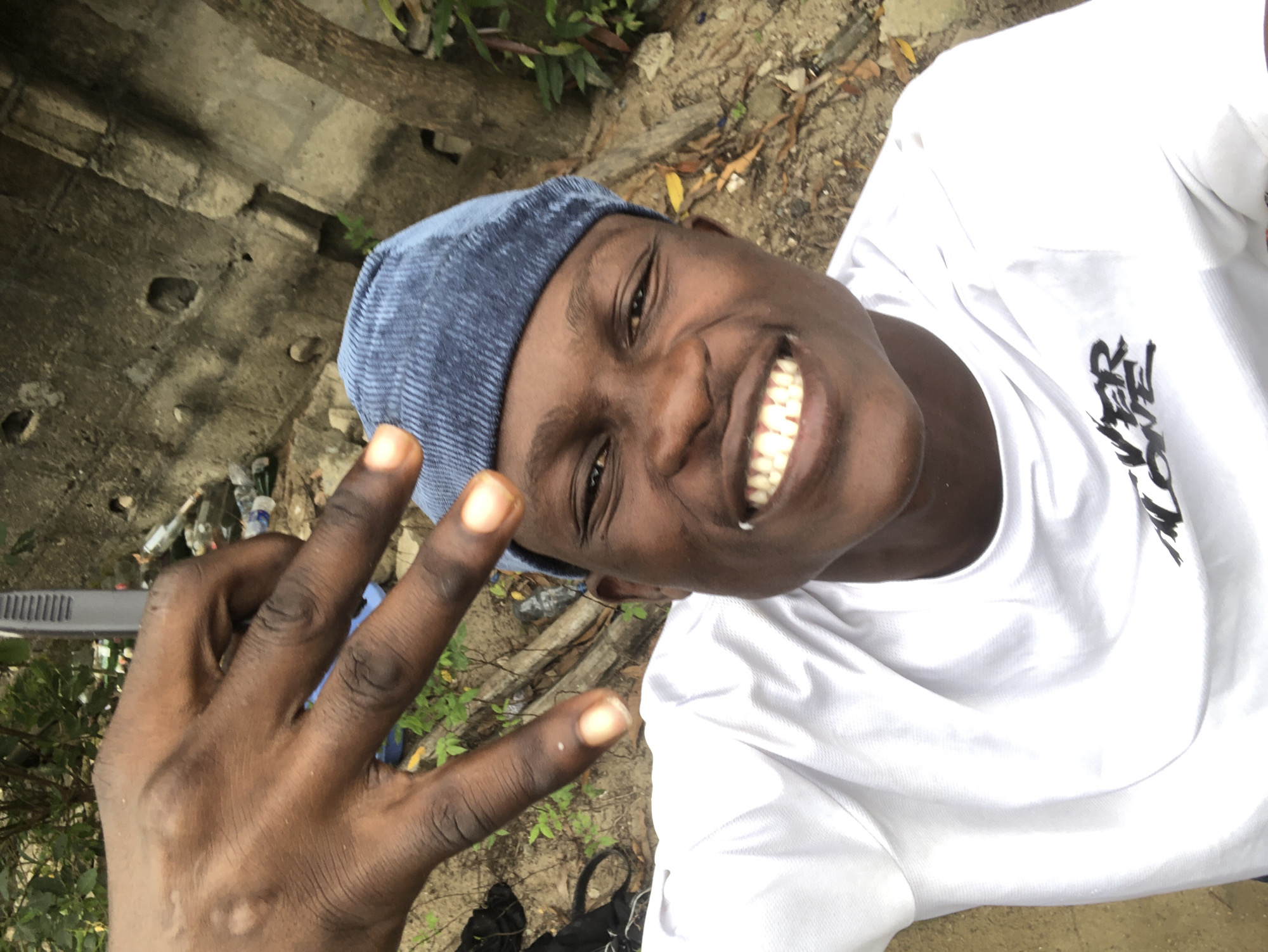In the city of Ado, Nigeria, a young Black male is captured in a close-up, eye-level, selfie-style photograph. He is wearing a blue, ribbed beanie and a white t-shirt with black lettering that reads "NEVER ALONE". His face is turned towards the camera, and he is smiling broadly, displaying his teeth. His right hand is raised, with the index and middle fingers extended in a peace sign gesture, positioned in the upper left quadrant of the frame. The background is out of focus, but appears to be an outdoor, somewhat unkempt environment with vegetation, a rough concrete wall, and scattered debris, including plastic bottles and what looks like a discarded electronic device. The lighting suggests it is daytime.