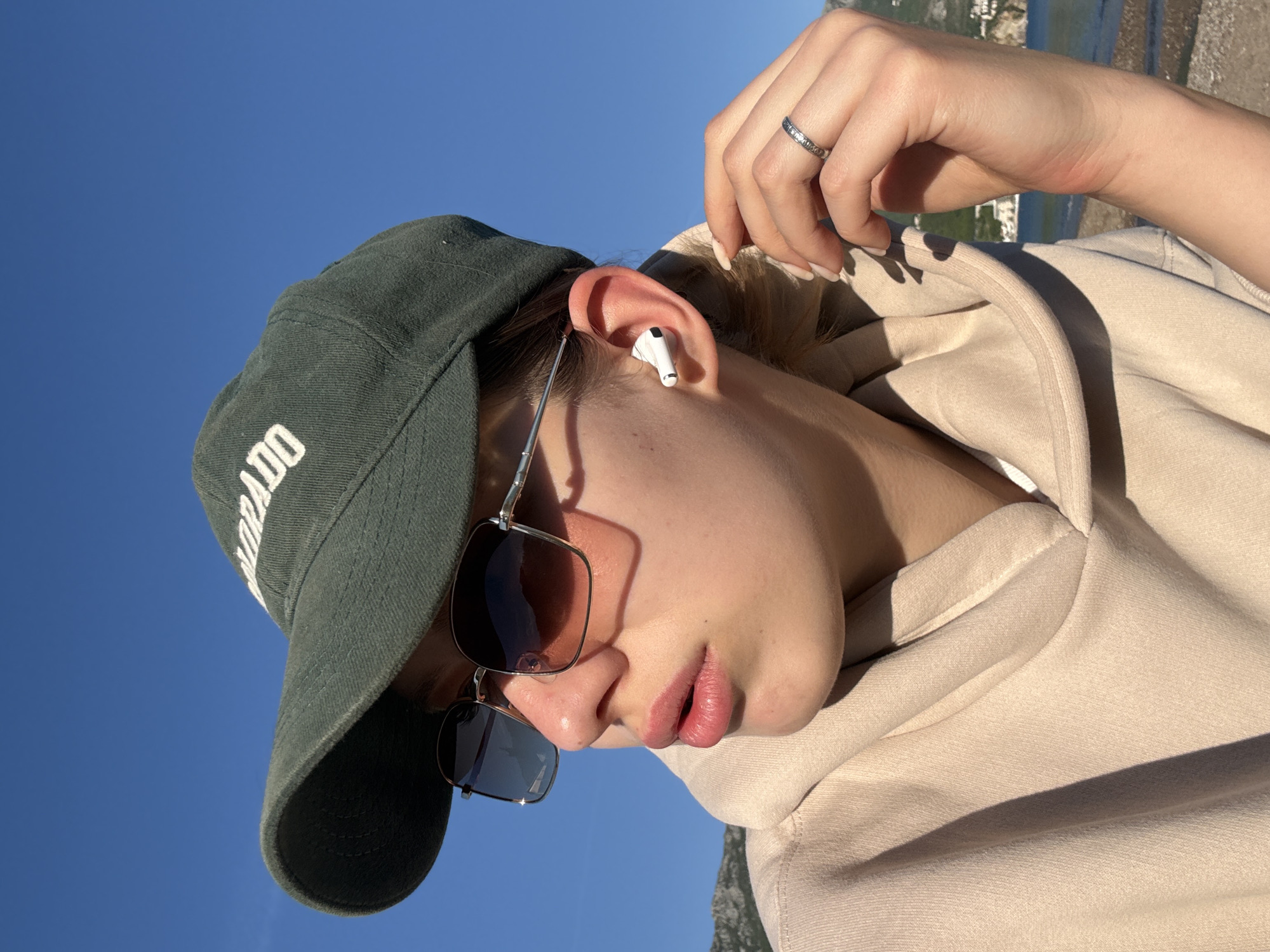 A close-up, angled portrait captures a person outdoors in Boreti, Montenegro, under a clear, bright blue sky. The subject, seen from the side, wears a dark green baseball cap with partially visible white text "ORADO" (likely "COLORADO"), square-framed sunglasses with dark lenses, and a light beige hooded sweatshirt. A white wireless earbud (AirPod) is inserted into their right ear. The person's right hand is raised towards their head, with a silver-colored ring featuring small inset stones on the ring finger. Their fingernails are neatly manicured with light polish.
The image is taken in strong, direct sunlight, casting distinct shadows on the face, under the cap brim, and across the hoodie, suggesting either morning or late afternoon. The sky is an intense, unblemished blue, indicating fair weather. In the blurred background, hints of a distant landscape with water and possibly mountains are visible, consistent with a coastal or lakeside setting. The person appears to be looking down or away, creating a relaxed, introspective mood. The primary activity depicted is listening to audio via the earbud.