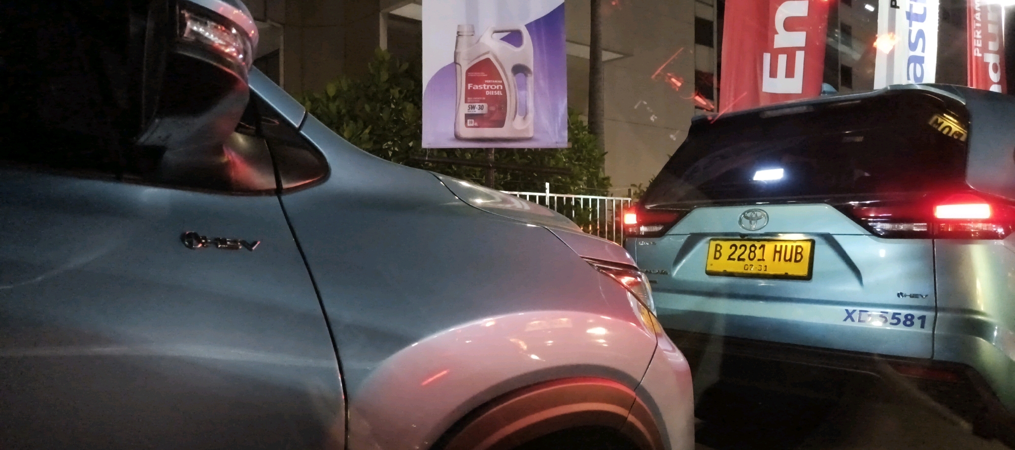 In the city of South Jakarta, Indonesia, at night, two blue cars are parked side-by-side. The car on the right, a Toyota, displays a yellow license plate with the characters "B 2281 HUB" and "07-31" below it. The rear of this car is visible, including its taillights, one of which is illuminated red. Below the license plate, a blue decal reads "XD5581". The car on the left, a different model, is partially visible, showing its side mirror and a chrome emblem reading "CHEV". Behind the cars, a large banner advertises "Fastron Diesel SW-30" engine oil. Trees and foliage are visible in the background, along with a white metal fence. The scene is illuminated by streetlights and the lights of the vehicles.