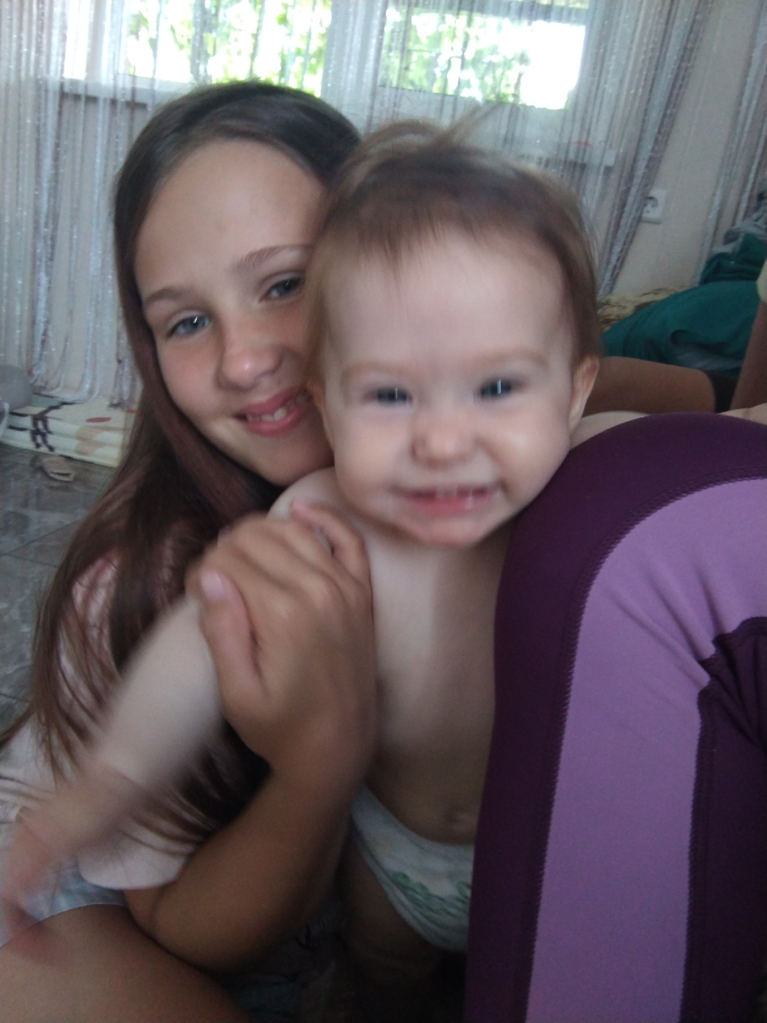 The media file shows a close-up, slightly blurry selfie of two young individuals. On the left, a girl with long, dark brown hair and blue eyes is smiling. She is holding a baby, who is in the foreground and also smiling, showing some teeth. The baby appears to be wearing a diaper with a green pattern. The girl is wearing a light-colored shirt. The person taking the photo is not fully visible, but a portion of their legs in purple leggings can be seen on the right side of the frame.
The background suggests an indoor setting. Behind the subjects, sheer white curtains are visible, with sunlight filtering through them, indicating it's likely daytime. Outside the window, greenery can be seen, hinting at a location with trees or plants. The floor appears to be tiled. The overall mood seems joyful and intimate, captured in a candid moment. There is no visible text in the image. The context provided places the location as Vinogradny, Russia.