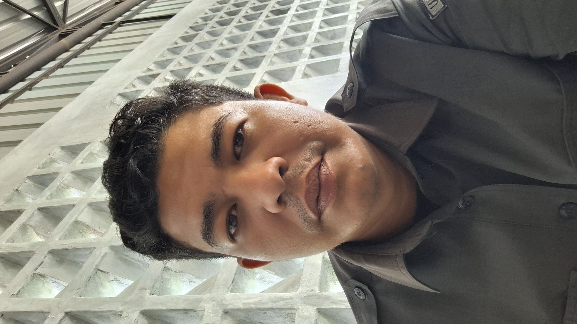 The image features a close-up, angled portrait of a man, likely a selfie. He is positioned prominently in the foreground, looking directly at the camera with a subtle, composed expression. He has short, dark, wavy hair, and his skin tone is medium brown with some visible stubble on his chin and upper lip, along with minor blemishes or acne scars on his left cheek. His eyes are dark.
The man is wearing a dark-colored collared shirt, possibly dark green or brown, with buttons visible down the front. A small, rectangular patch or emblem with a white "U" symbol is visible on the upper left side of his shirt, near the shoulder.
The background consists of architectural elements. Behind the man, a light-colored wall with a prominent, repeating geometric pattern of recessed square or diamond shapes creates a textured grid. This suggests a decorative concrete block wall or similar architectural feature. Above this wall, towards the top left of the frame, a darker, angular roof structure with exposed beams or trusses is visible, indicating an indoor or covered outdoor setting, possibly a modern building, cafe, or public space in Lhokseumawe, Indonesia. The lighting appears soft and diffused, suggesting daytime or well-lit interior conditions. No specific activity or event other than being photographed is depicted.