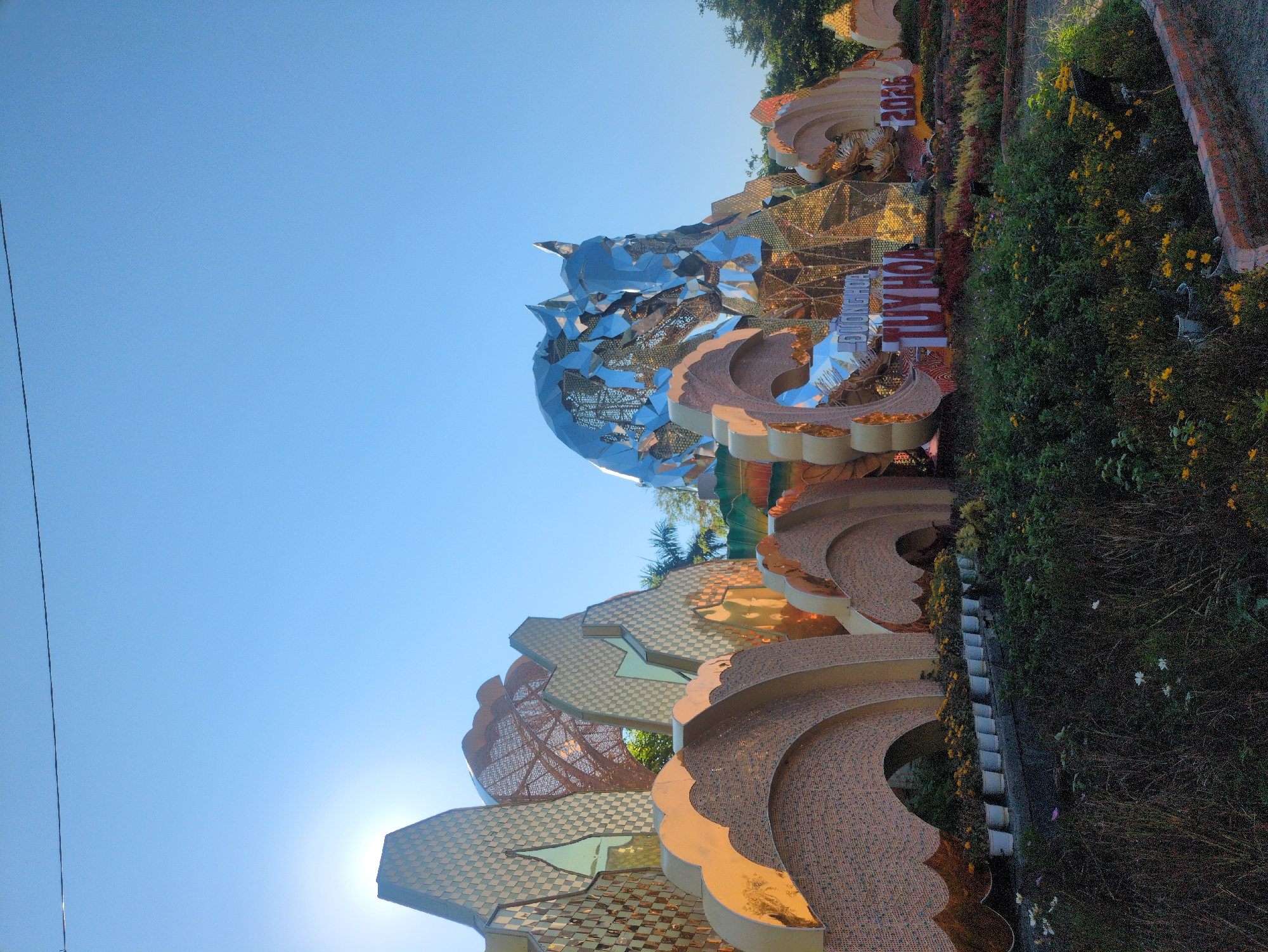 The image depicts a large, elaborate sculpture in Tuy Hòa, Vietnam. The sculpture is comprised of curving, tiered structures covered in mosaic patterns and topped with a fractured, mirrored sphere. It is partially surrounded by vegetation and flowers, with the inscription "TUY HOA" visible. The sky is clear and blue. A brick wall is on the right side of the image.
