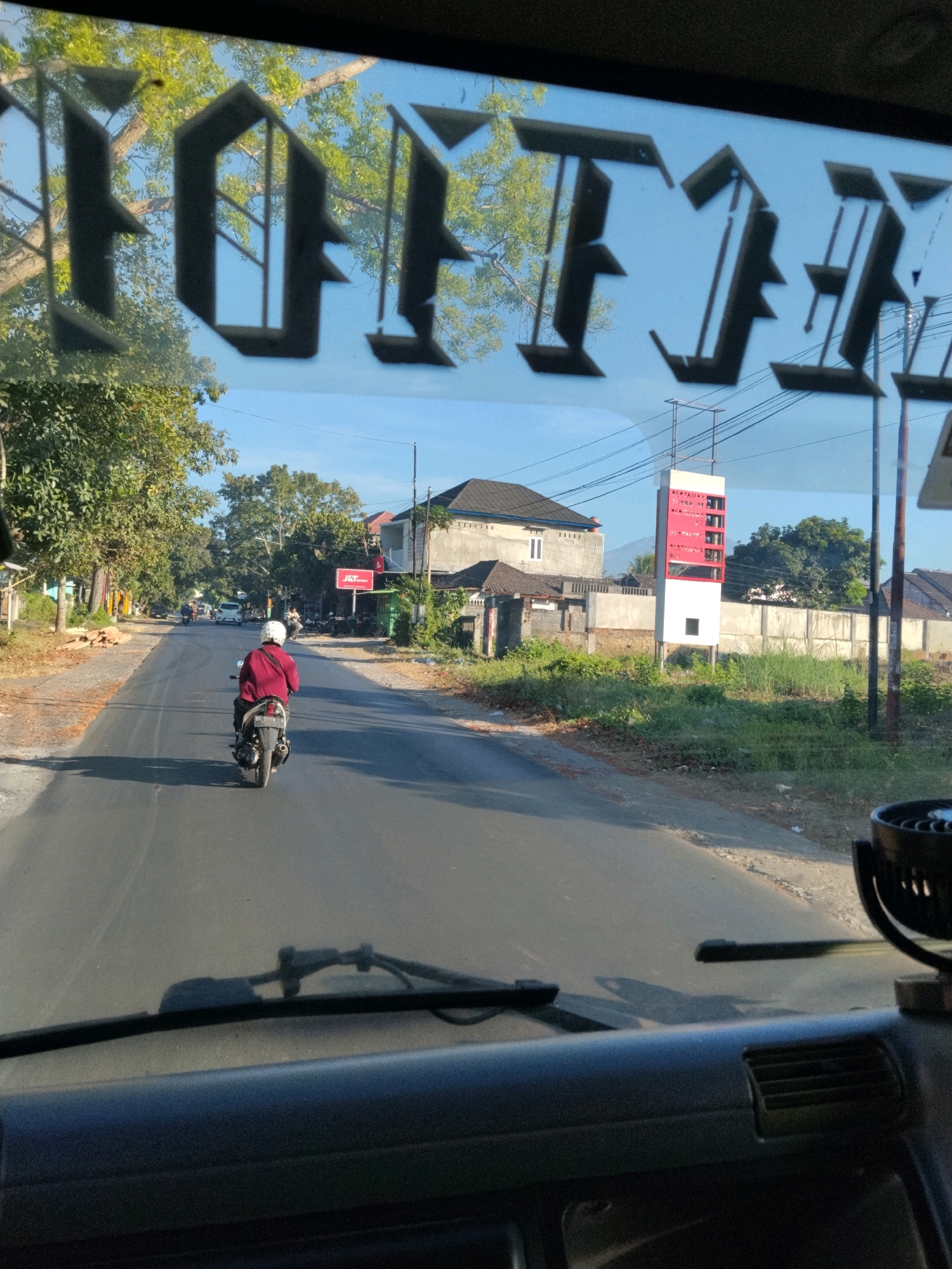The image is taken from inside a vehicle, looking forward through the windshield. The windshield has decorative black lettering applied to it, partially obscuring the view of the road ahead. The lettering appears to be in a gothic style and spells out "OLTIE."
The scene outside depicts a rural or suburban road in Kelayu Utara, Indonesia. The road is paved and appears to be two lanes. A person is riding a motorcycle in the right lane, facing away from the camera, wearing a red jacket and a white helmet. Trees line the sides of the road, and there are buildings visible in the background, including what appears to be a house and a commercial building with a red and white sign that says "PERTAMINA" and "PERTAMAX." There are also power lines and utility poles. The sky is clear and blue. The overall lighting suggests it is daytime, likely morning or afternoon, with the sun casting shadows.