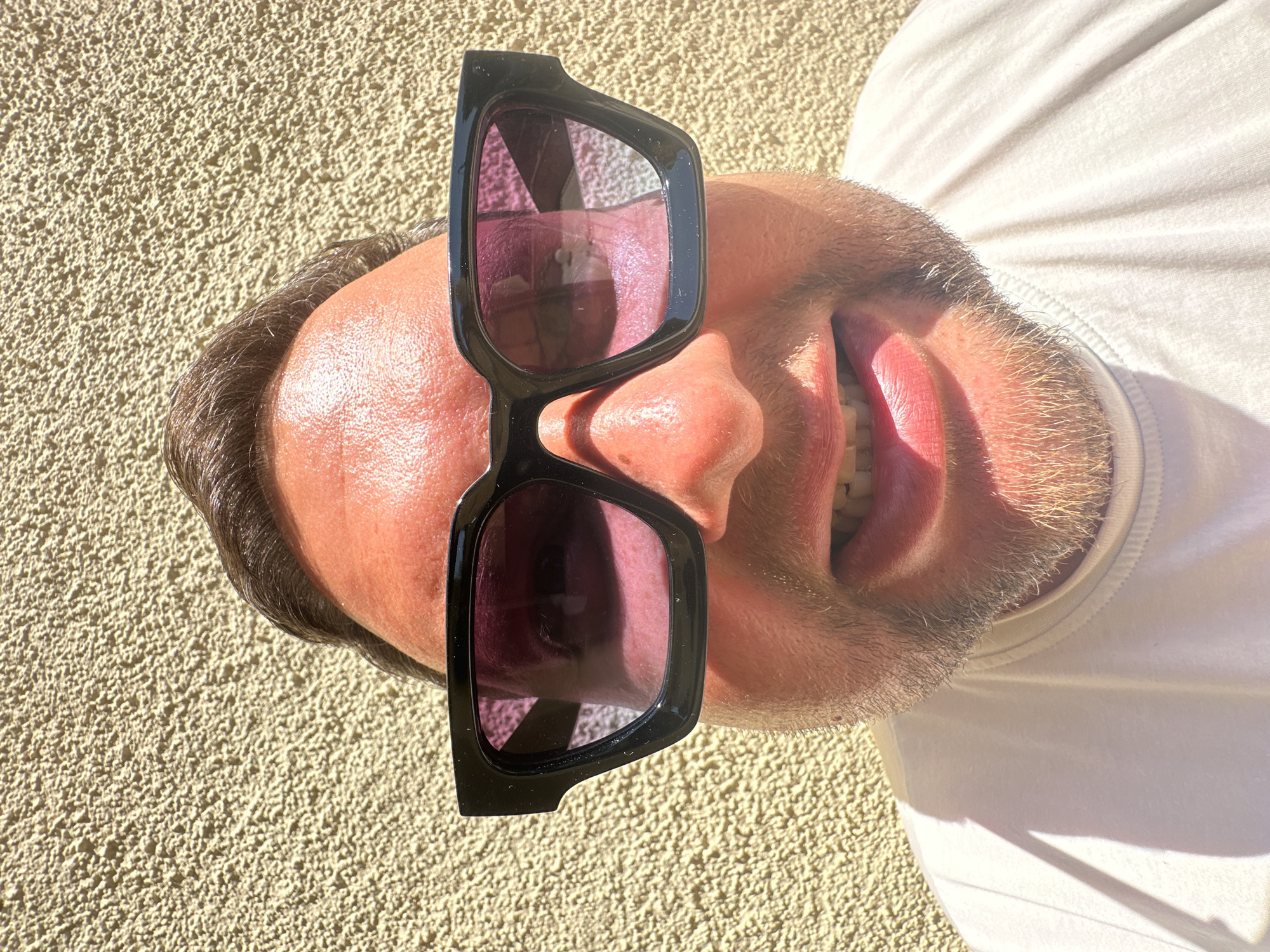 A close-up, angled shot depicts a man with fair skin, a beard, and short brown hair, wearing a white t-shirt. He is also wearing large, black-rimmed sunglasses with purple-tinted lenses, which appear to be resting on his nose. He is smiling, exposing his upper teeth. The background consists of a textured, light-yellow stucco wall.
There is no evidence of location in Sant Feliu de Guíxols, Spain.
