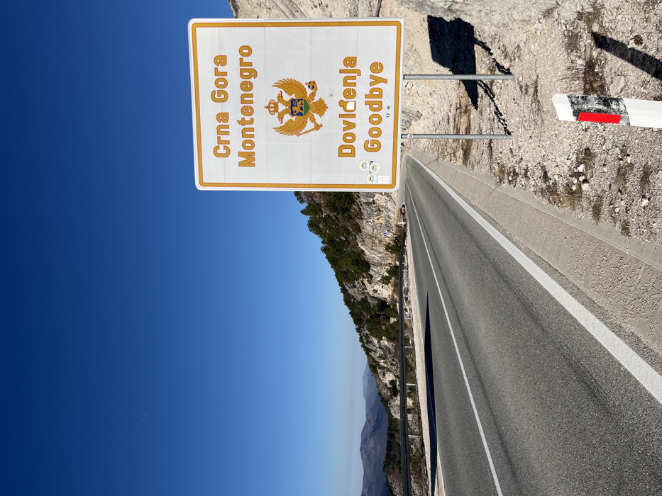 The image depicts a mountainous border area near Klobuk, Bosnia and Herzegovina, under a clear blue sky. In the foreground, a prominent white rectangular road sign with an orange border is visible on the right side of an asphalt road. The sign, supported by two grey metal posts, displays "Crna Gora Montenegro" at the top, followed by the gold Montenegrin coat of arms (a double-headed eagle holding an orb and scepter with a lion on a shield), and "Doviđenja Goodbye" at the bottom. Several small circular stickers are affixed to the lower left portion of the sign. To the immediate right of the sign's base, a white and black reflector post with a vertical red stripe is partially visible in the gravel.
The two-lane asphalt road, marked with solid white lines, curves slightly to the left and ascends into the distance. A concrete curb separates the road from a gravel and rocky shoulder on the right. On the left side of the road, a metal guardrail runs parallel to a steep, rocky mountainside partially covered with green pine trees and sparse vegetation. Distant mountains are visible on the horizon. The scene is brightly illuminated by direct sunlight, casting distinct shadows to the right of the sign and on the left side of the road. A faint airplane contrail is visible in the upper left quadrant of the sky. No people or vehicles are present in the frame.