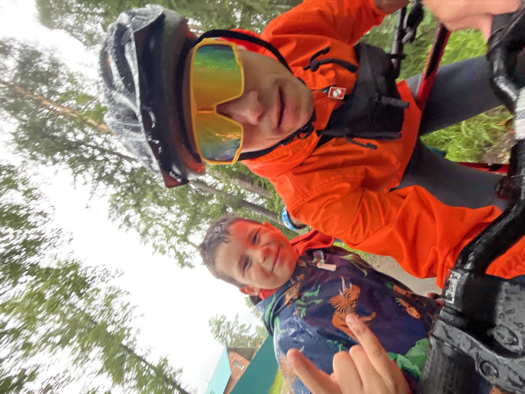 This is a selfie taken by two people on bicycles in Chaikovsky, Russia. The person in the foreground is a child wearing a blue raincoat with a dinosaur pattern. They are smiling and pointing towards the camera. The person behind them is wearing an orange jacket and a black helmet, with large, reflective sunglasses that obscure their eyes. The scene appears to be outdoors, with trees and greenery visible in the background. The ground is wet, suggesting it might be raining or has recently rained. The sky is overcast, indicating a cloudy day. The overall mood of the image is cheerful and adventurous, with the subjects enjoying a bike ride together.