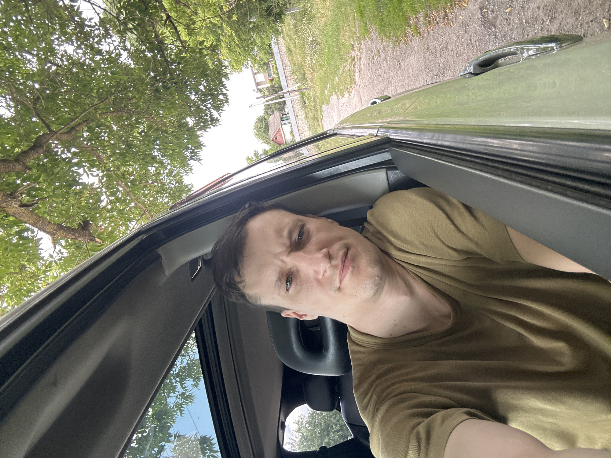 A man is taking a selfie from the passenger seat of a car. He is wearing a tan t-shirt and looking at the camera with a slightly puzzled expression. The car has a sunroof that is open, and sunlight is filtering through the trees above. The car is parked on a dirt road next to a grassy field with some houses in the background. The location is identified as Makiv, Ukraine.