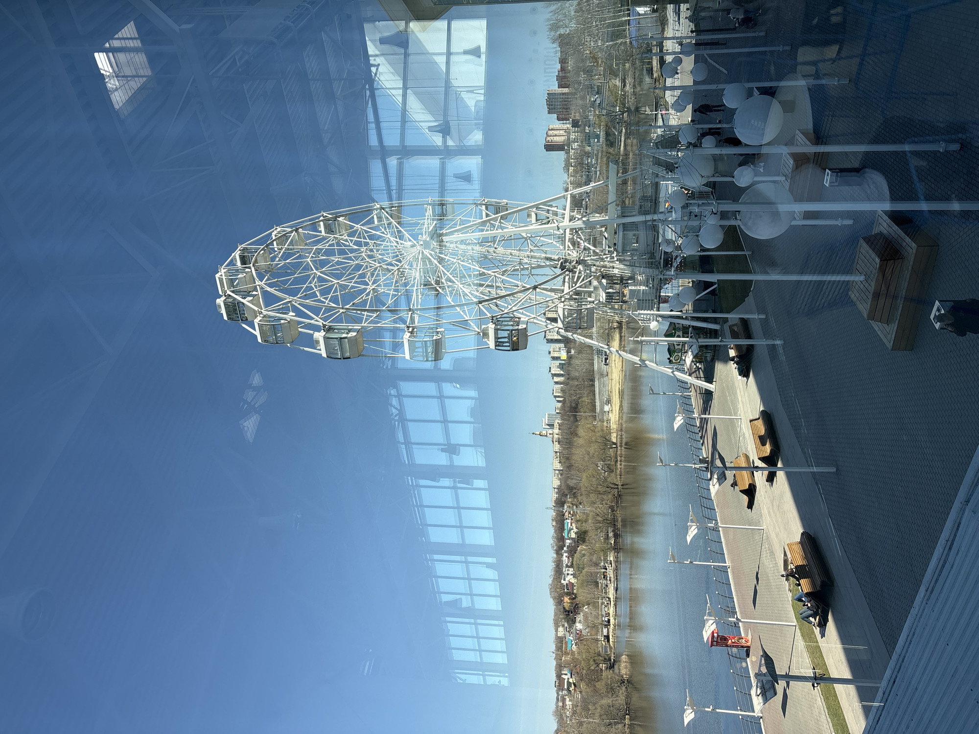 The image depicts a view, possibly from a window, overlooking the city of Ulyanovsk, Russia. A prominent Ferris wheel is visible, along with the Volga river and a paved promenade featuring benches and lampposts. The image is likely taken during the day, with clear blue skies. The perspective is somewhat distorted due to the window reflection, which also shows a building's structure.