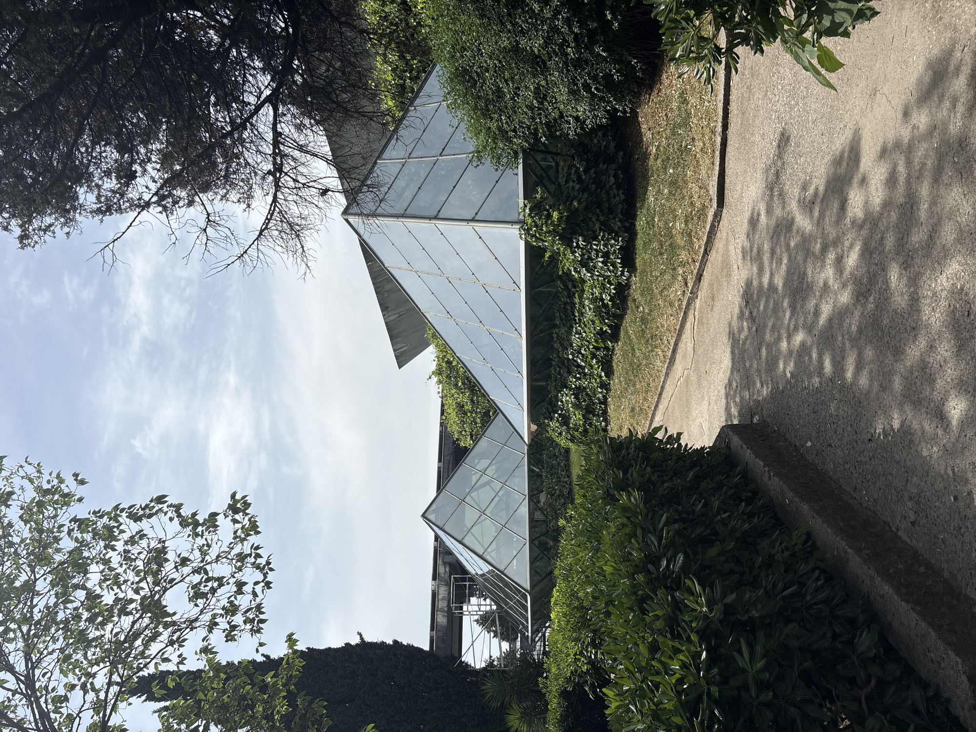 The image shows two glass pyramid structures located in a park or garden setting. Lush green bushes and trees surround the pyramids. A concrete walkway borders the greenery. The sky is visible through the trees, with scattered clouds. The pyramids are modern architecture, constructed from clear glass panes and a supporting framework. Scaffolding can be seen behind one of the pyramids. The location is Podgorica, Montenegro.