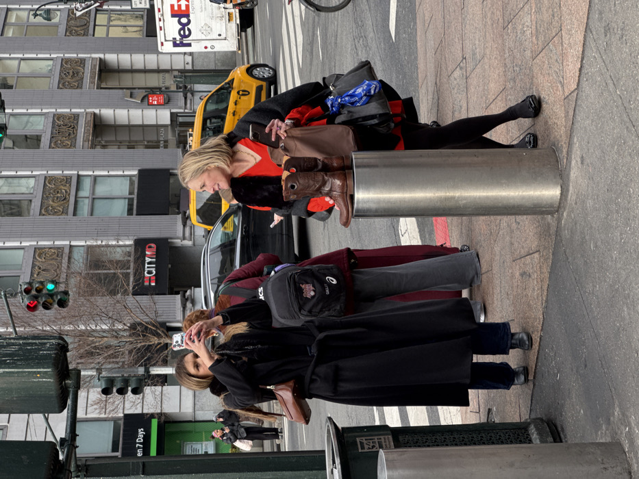 A group of women gathers on a city sidewalk, one taking a photo with her phone. Another woman with blond hair poses with cowboy boots atop a metal bollard, using her phone to take a selfie. A yellow taxi and a FedEx truck can be seen in the background alongside tall buildings. The day appears bright and sunny.