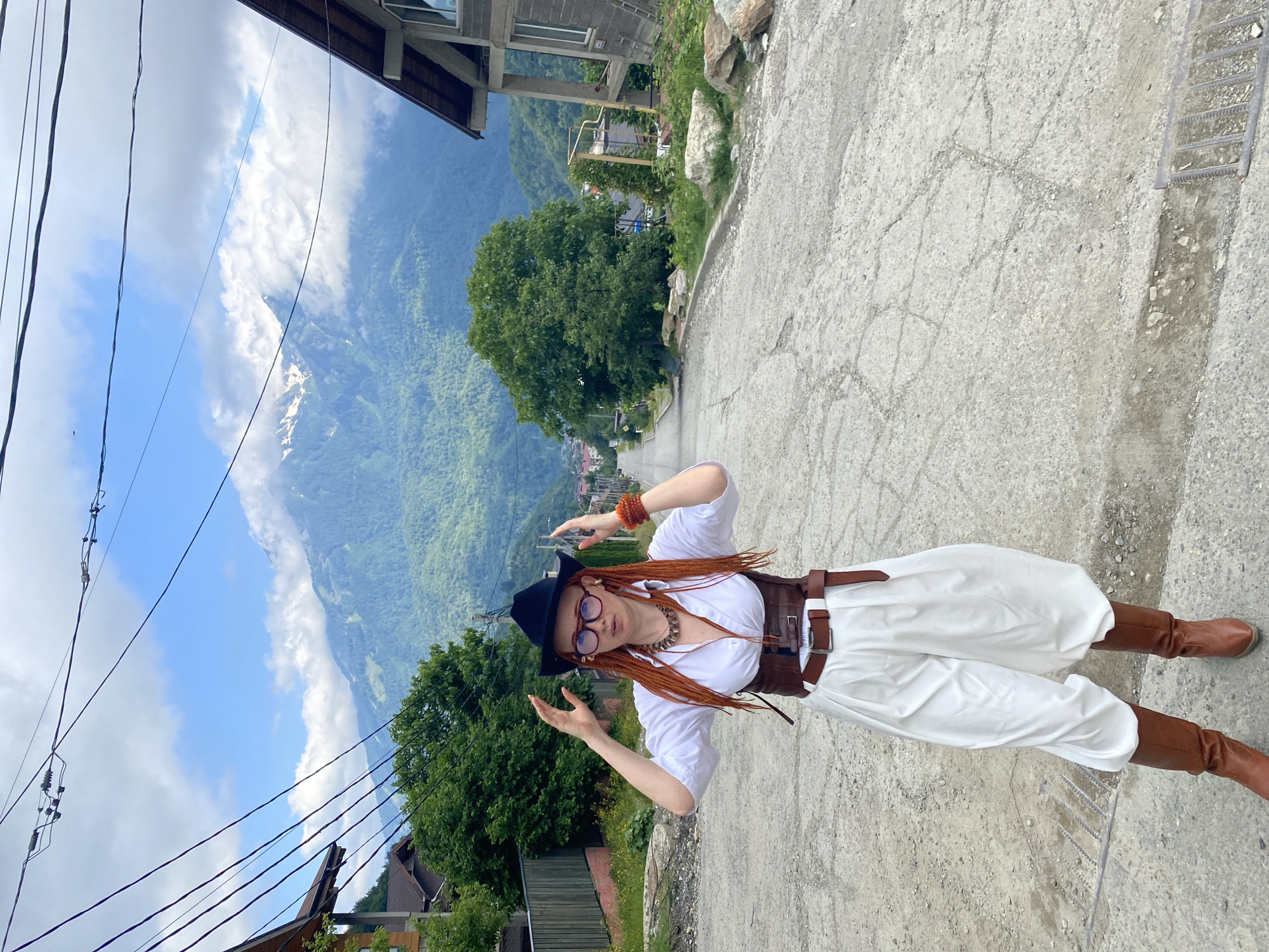 In Krasnaya Polyana, Russia, a woman stands on a steep, paved road with cracked asphalt. She is wearing a black cowboy hat, large-rimmed glasses, a white shirt, a brown corset, white billowing pants, and brown knee-high boots. Her orange braided hair is visible beneath her hat. She has her hands raised to her head, fingers spread. In the background, lush green mountains are visible, partially obscured by clouds. Homes are clustered along the hillside. Utility lines are strung across the sky.