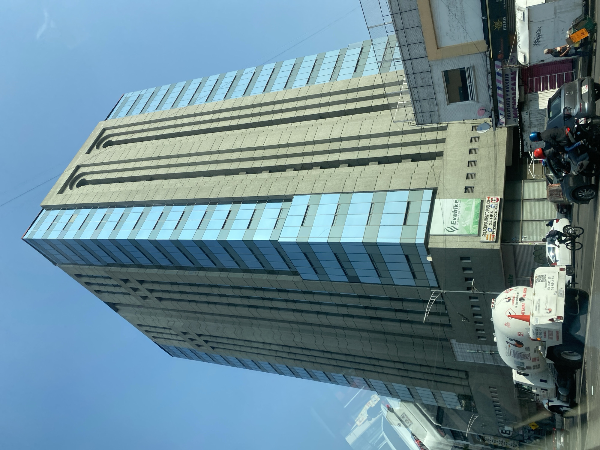 The image captures a tall building with alternating bands of glass windows and concrete panels, set against a clear blue sky. The building occupies a significant portion of the frame, dwarfing the surrounding street-level scene.
On the right side of the building, a smaller section with fewer windows rises. In front of this section, there's a street scene, where multiple vehicles, including a white truck with a tank-like structure and a motorcycle with two riders, are parked. A sign for "Evobike" is visible on the building. Other commercial signs are also visible above the street-level shops and businesses.
The overall environment suggests a busy urban area, possibly a commercial district.