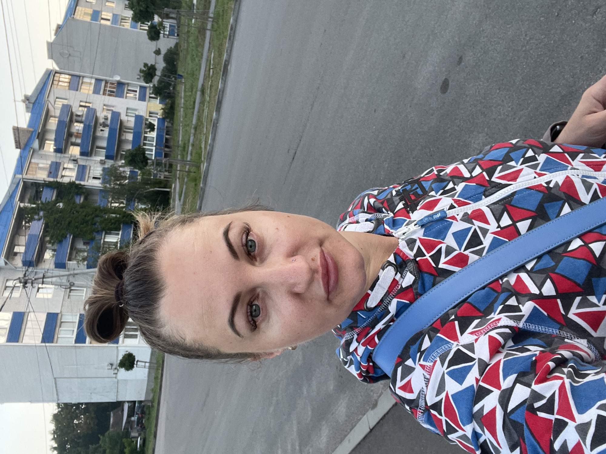 A mid-shot, outdoor selfie captures a woman standing on a paved road in front of apartment buildings. The woman, positioned on the left side of the frame, has her hair styled in a top knot and wears a colorful hooded jacket with a geometric pattern of red, blue, and black triangles on a white background. A blue strap from a bag crosses her chest. Her facial features are visible, including her eyes, nose, and lips. Behind her, several multi-story apartment buildings with blue balconies and white windows line a street. Trees are visible in front of the buildings, and power lines are strung between poles. The road surface is asphalt, with a sidewalk or curb on the far left. The sky is overcast, suggesting it is daytime. This scene is located in the city of Zarichne, Ukraine.