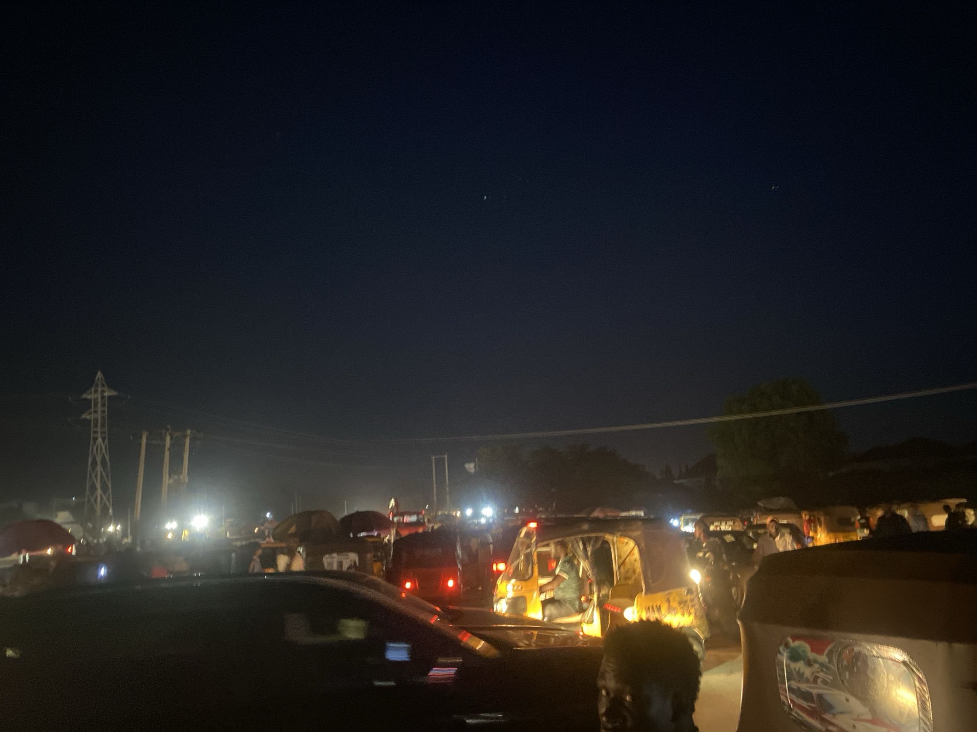 The image shows a night scene in Abuja, Nigeria, possibly a street or market area based on the presence of tuk-tuks (auto rickshaws), umbrellas (likely market stalls), and overhead power lines/electrical poles. People are visible, though details are obscured by darkness and distance. The scene is illuminated by streetlights and vehicle headlights. The sky is dark, with a few visible stars.