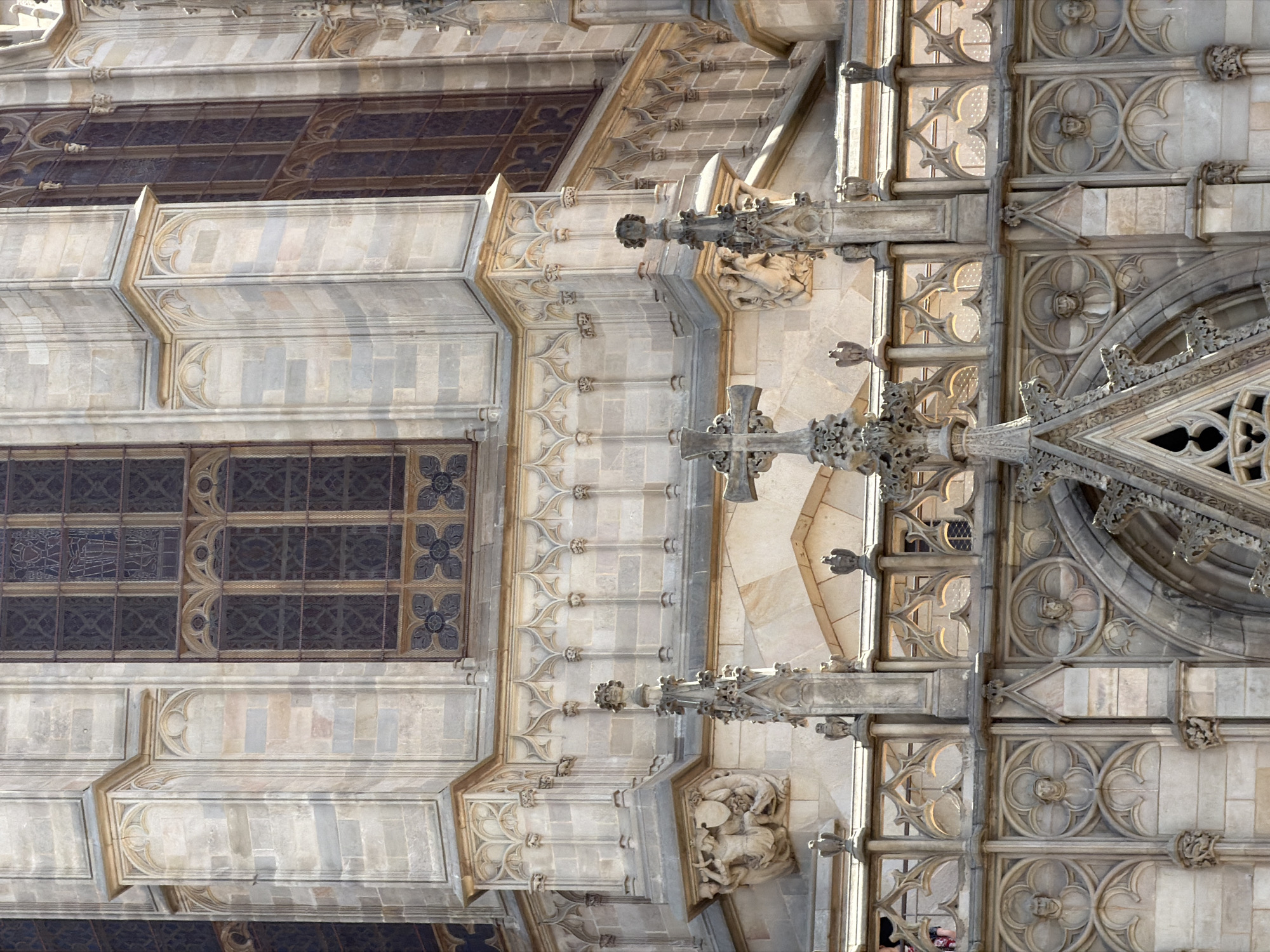 The image shows a close-up, eye-level view of the ornate facade of a Gothic cathedral in Barcelona, Spain. The architecture features intricate stone carvings, detailed tracery in the windows, and decorative elements such as gargoyles and sculpted figures. The sunlight highlights the texture of the stone, casting shadows that accentuate the three-dimensional nature of the carvings. The windows are tall and narrow, filled with dark stained glass. A prominent cross is visible in the foreground, and further back, decorative railings and sculpted medallions depicting faces are integrated into the facade. The building appears to be made of light-colored stone, with some sections showing variations in color due to age or material. The overall impression is one of historical grandeur and meticulous craftsmanship.