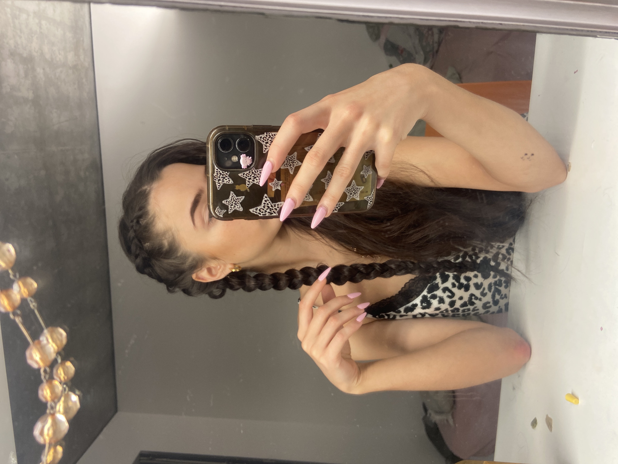 A woman is taking a selfie in a mirror. She has long, dark hair styled in braids. Her nails are long and painted light pink. She is wearing a top with a leopard print pattern and black lace trim. Her phone has a patterned case with stars and a small pink object attached. The background shows a mirror, a dresser, and a small tattoo on the woman's arm. The location is not identifiable from the image.