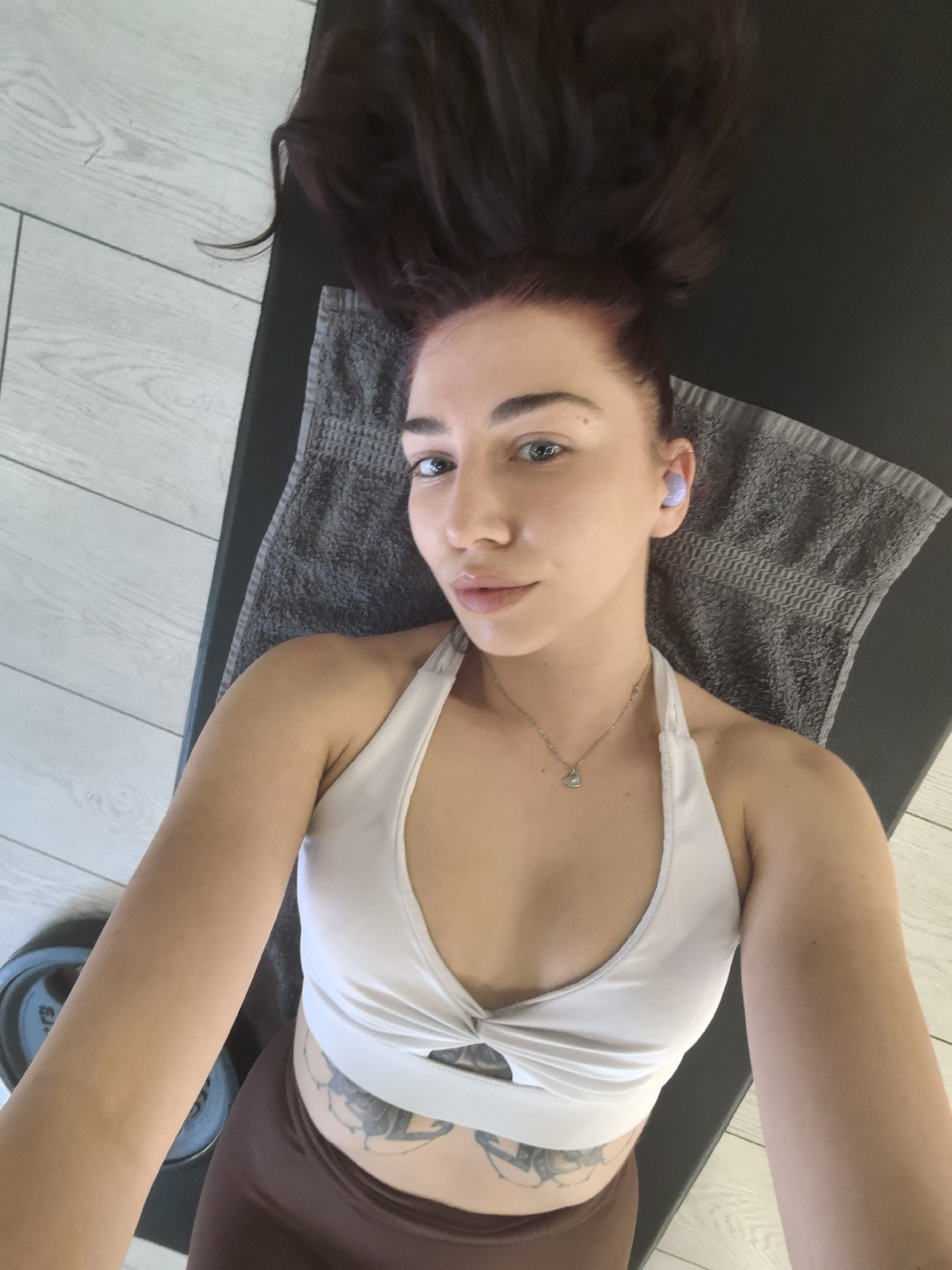 A person is lying on a yoga mat. A grey towel is beneath their head. The person is wearing a white halter top and brown leggings. Tattoos are visible on their abdomen. They are also wearing light purple earbuds. Two weight plates are visible to the left. The location is a room with light grey wood-look flooring.