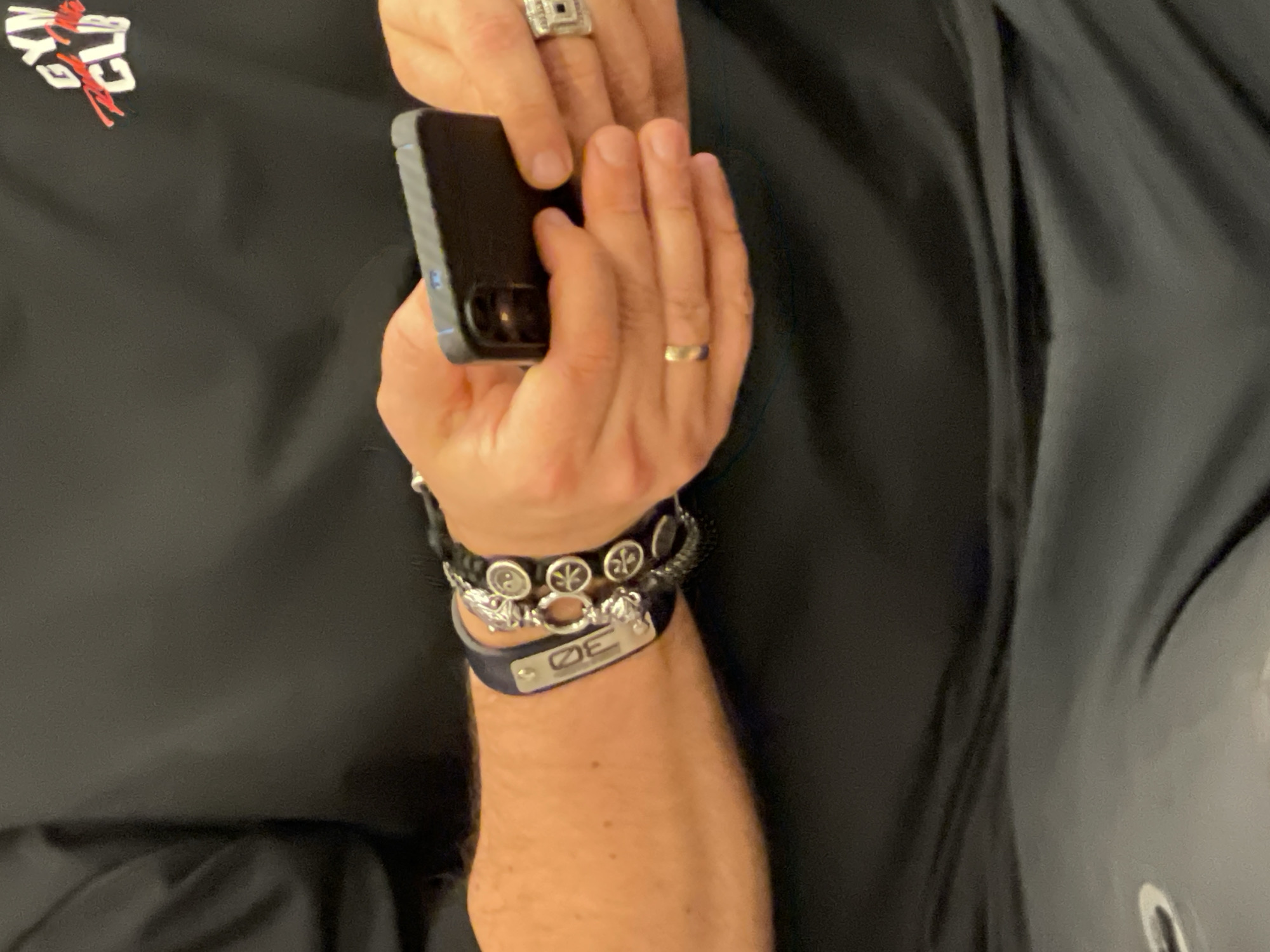The image depicts a close-up view of a person's upper torso, arms, and hands, actively engaging with a smartphone in an indoor setting in Moscow, Russia. The individual, whose gender is not explicitly discernible but appears masculine, wears a black short-sleeved top. A partial logo on the upper right chest area of the top reads "GYM CLUB" in white block letters, with a red stylized script appearing between the two words. The lower left portion of the frame shows blurred dark grey or black athletic wear, possibly with a faint white or silver logo.
The person's left forearm is visible, adorned with multiple bracelets. A prominent black band, likely leather or silicone, features a rectangular silver-colored metal plate engraved with two stylized characters, resembling "SO" or "ЭО". Adjacent to this, a multi-strand bracelet, possibly beaded or braided, displays silver-colored charms including a stylized skull, a ring-like connector, and several circular elements with a cross-like pattern. On the left hand's ring finger, a large silver-colored ring with a geometric, rectangular top is worn.
Both hands are occupied holding a dark-colored smartphone, which has a dark protective case and shows a visible camera module on its rear. The thumbs are positioned as if actively interacting with the device. A plain gold-colored band ring is worn on the fourth finger of the right hand. The background is indistinct and dark, offering no further environmental details.