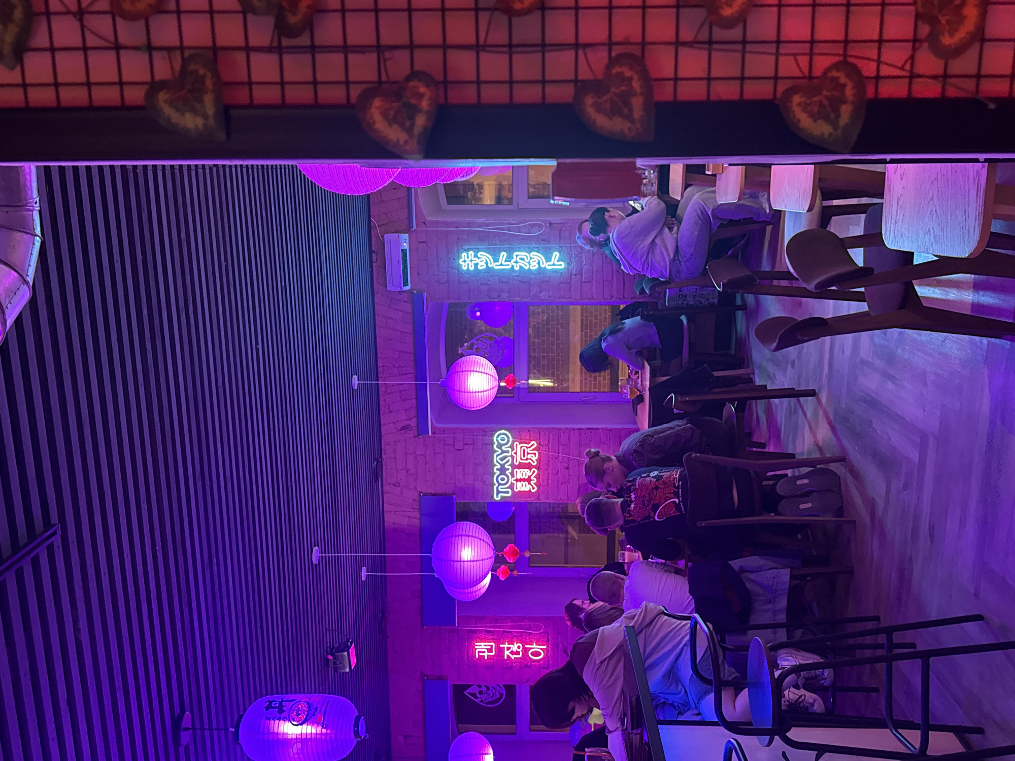 Here is a description of the image:
The image shows the interior of a restaurant illuminated with neon lights, giving off a purple and pink hue. People are seated at tables, appearing to be engaged in conversations or eating. There are hanging paper lanterns, contributing to the overall atmosphere. Neon signs with text in foreign languages (presumably Japanese) are visible on the brick walls. The restaurant has a modern and somewhat stylized decor, with wooden furniture and a distinctive grid-like design on the ceiling. The scene suggests a lively, urban dining environment.
