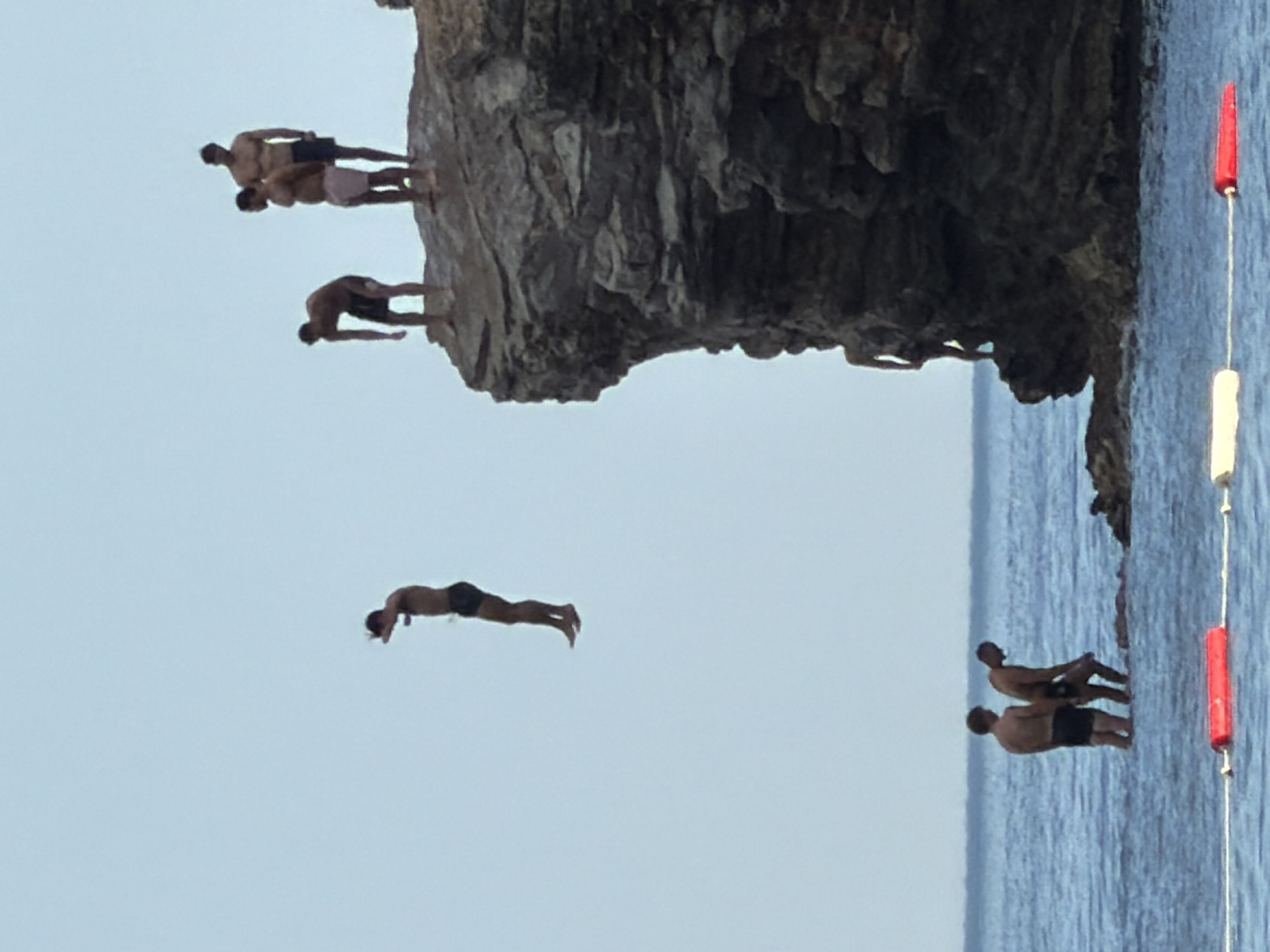 The image depicts a group of people engaging in cliff diving in Budva, Montenegro. Several individuals are visible on a rocky cliff face, some poised to jump, while others are in mid-air. One person is captured in a dynamic diving pose, arms extended and body streamlined. Below, the clear blue water of the Adriatic Sea stretches out, inviting for a plunge. Two figures stand on the edge of the water, possibly waiting for their turn or observing the divers. The scene is illuminated by what appears to be daylight, casting soft shadows and highlighting the rugged texture of the rocks. The clear sky suggests fair weather conditions. The overall atmosphere is one of adventure and exhilaration, as the individuals take part in a thrilling recreational activity. The presence of buoys with red and white floats in the water indicates a designated swimming or activity area.