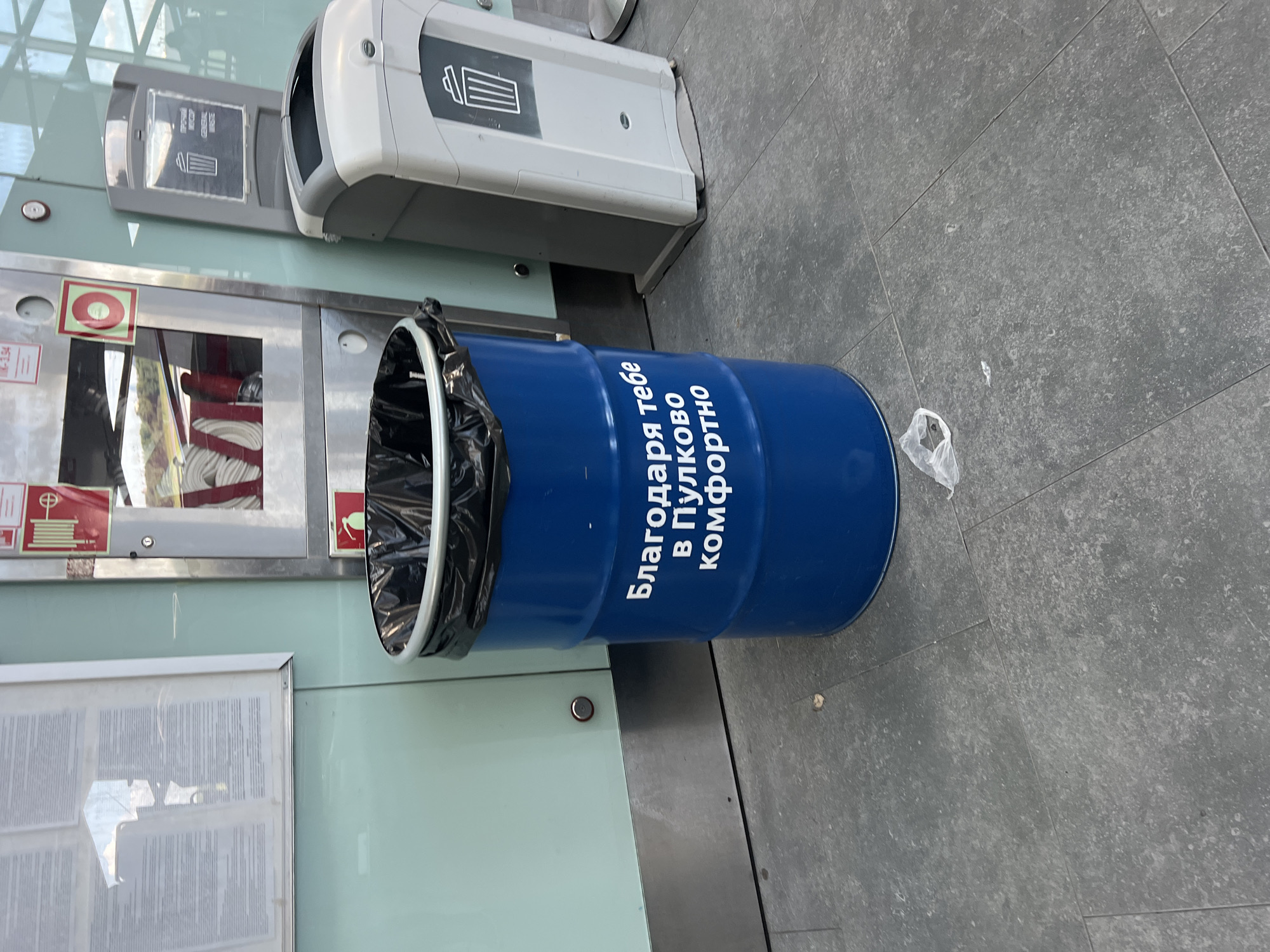 This is a medium-shot, eye-level, full-body shot of a blue cylindrical trash can with white lettering. The trash can is in the foreground, with a black plastic bag lining the top. The white lettering on the trash can reads "Благодаря тебе в Пулково комфортно," which translates to "Thank you for the comfort in Pulkovo." The trash can is positioned on a gray tiled floor. To the right of the trash can is a white and gray public waste bin. In the background, there is a wall with light blue panels and a metal base. There are also some informational displays on the wall and a large metal cabinet with red and white signage. The scene appears to be in an indoor public space, possibly an airport terminal or a train station in the city of Saint Petersburg, Russia, given the reference to "Pulkovo," which is the name of Pulkovo Airport in St. Petersburg. There are no people visible in the image.