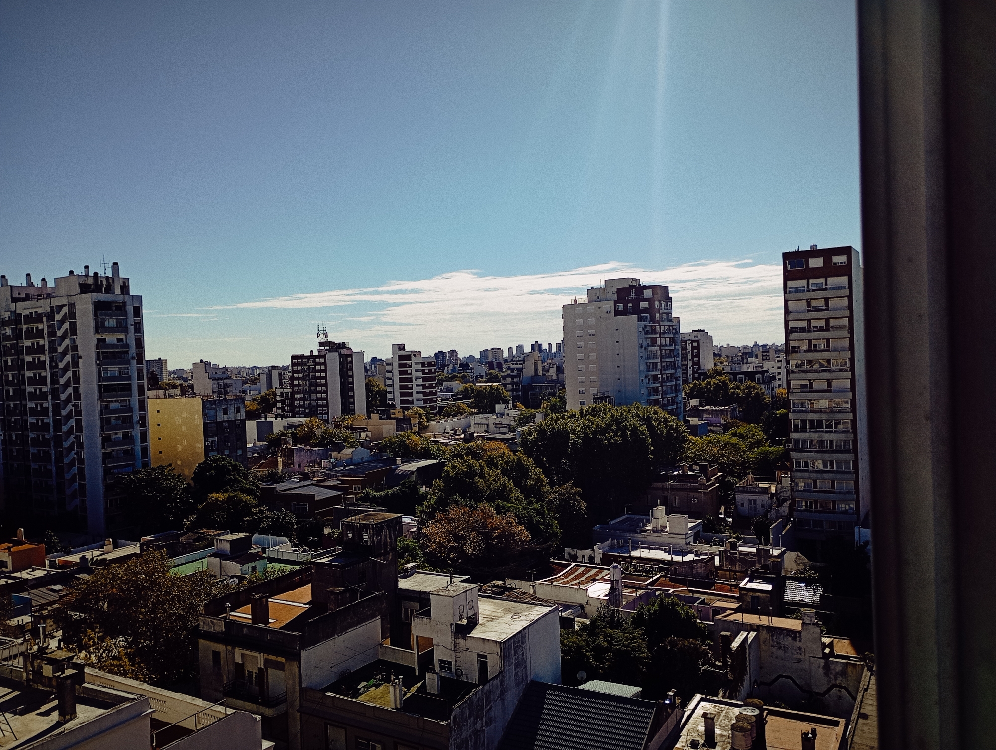 The image captures an expansive, elevated view of an urban landscape in Buenos Aires, Argentina, under a bright, clear sky. The perspective is from a high-rise building, looking out over a dense mix of architectural styles.
The upper third of the frame is a vivid blue sky, mostly clear with a few scattered, wispy white clouds primarily along the horizon. A strong lens flare in the upper right corner indicates direct and intense sunlight, suggesting the photo was taken during mid-day to early afternoon on a clear, sunny day.
The city below is characterized by a blend of tall, modern apartment buildings and older, lower-rise structures. On the far left, a prominent multi-story residential building with numerous balconies stands tall. Towards the center-right, another significant apartment building with lighter facades and darker upper floors is visible. A slender, dark-windowed building rises on the far right, partially obscured by a dark vertical element, likely a window frame. The foreground and mid-ground reveal many smaller buildings with flat roofs, some featuring terracotta tiles, while others are grey or green, often showing rooftop utilities like water tanks.
A striking feature is the abundant presence of large, mature trees with dense foliage, interspersed throughout the urban fabric. While most trees are a vibrant green, some display rich autumnal hues of brown and orange, particularly in the mid-ground, adding color variation to the scene.
No people are visible, and there is no discernible text on any of the buildings or signs. The image portrays a tranquil yet bustling cityscape on a beautiful, sunlit day.