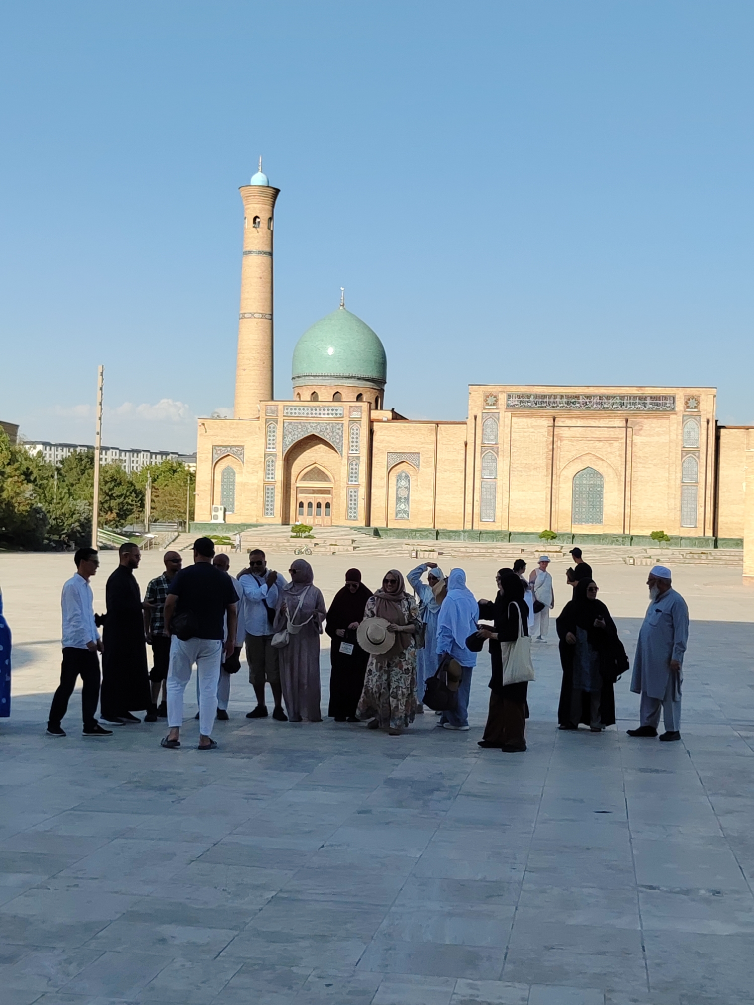 In Tashkent, Uzbekistan, a group of people is gathered in an open, paved courtyard in front of a large, ornate mosque. The mosque features a prominent minaret and a large turquoise dome, constructed from light-colored brick with intricate tilework. The people, appearing to be tourists and locals, are dressed in a mix of casual clothing and traditional attire, including headscarves and long robes. Some individuals are standing in conversation, while others are observing the mosque. The sky above is clear and blue, indicating a sunny day. Trees and buildings are visible in the background, suggesting an urban setting. The overall scene conveys a sense of cultural and architectural significance.