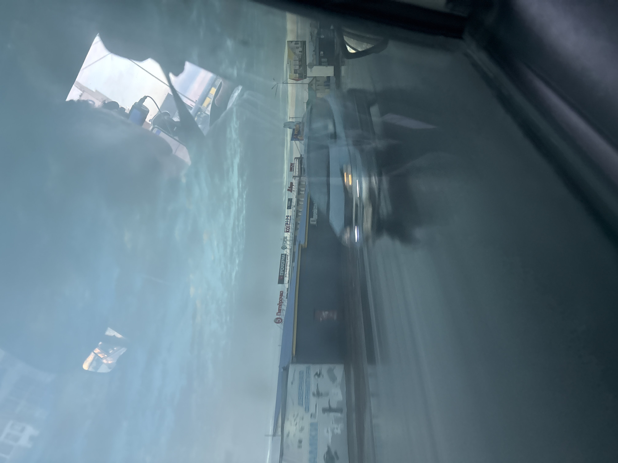 The image is taken from inside a vehicle, looking out through a windshield, and is oriented vertically as if rotated 90 degrees counter-clockwise.
The foreground features significant reflections on the windshield. On the left side, reflections show parts of the vehicle's interior, including what appears to be a blue object, a power adapter, and cables. On the lower left, there's a faint reflection of a grid pattern.
Outside the vehicle, a cloudy or overcast sky is visible, suggesting either a dull day, early morning, or late afternoon. A road or street extends into the distance, along which a dark-colored car with illuminated headlights is visible on the right side, possibly moving or stationary. The road surface appears somewhat reflective, suggesting it might be wet or recently rained on.
On the left side of the road, a commercial building or strip mall is present, featuring several signs. The most prominent legible sign is a red logo with white text for "Пятёрочка" (Pyaterochka), a well-known Russian supermarket chain. Above it, another sign reads "ПРОДУКТЫ" (Produkty), which means "Groceries" or "Food Products" in Russian. Other signs with Cyrillic text are visible but less clear. The presence of these Russian texts supports the provided location context of Chaikovsky, Russia.
No people are clearly visible, and the scene depicts a typical roadside environment. The blur of the moving car's lights could indicate motion or a slightly longer exposure.