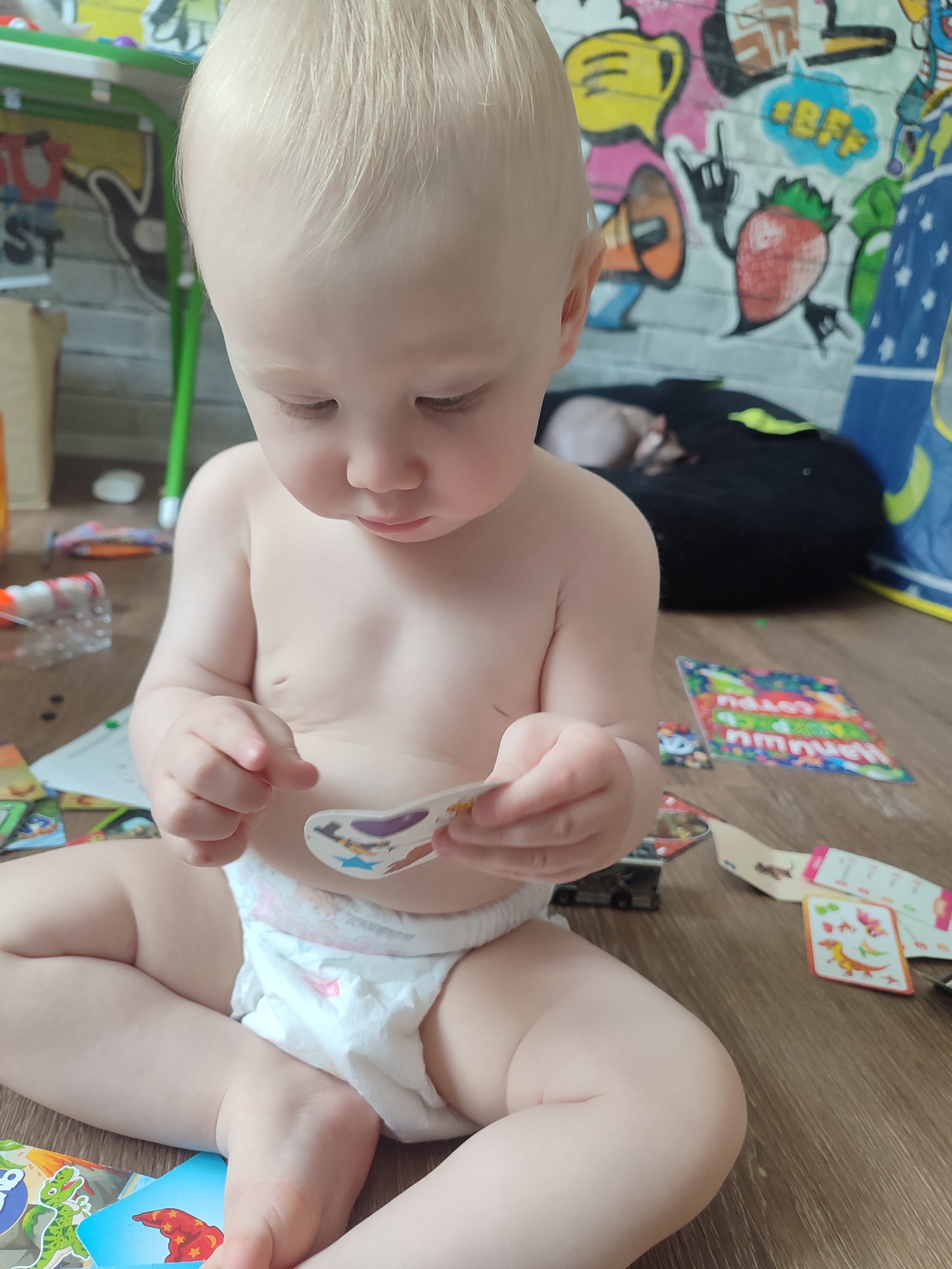 A baby wearing a diaper sits cross-legged on a wooden floor, holding a sticker in their hands. Scattered around the baby are various cards and toys. In the background, there is a colorful graffiti-style mural on a wall and a beanbag chair. The image was taken indoors, likely in a home environment. There is no definitive indication of the location in the city of Izhevsk, Russia, beyond the possibility of the location of where the image might have originated.