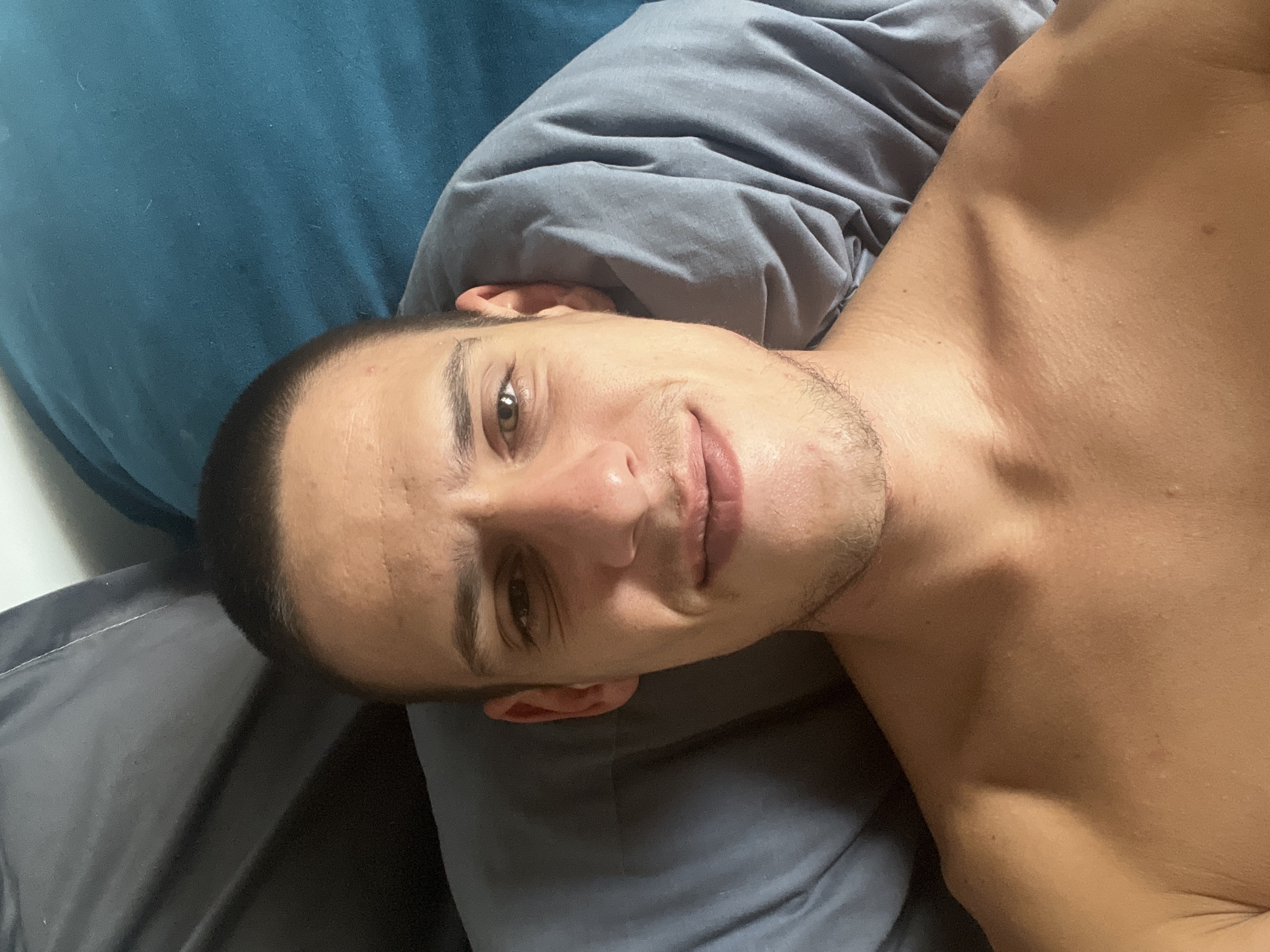 A shirtless man is lying on a bed with a blue pillow and a gray pillow. He is looking at the camera with a slight smile. The background is out of focus. No evidence to suggest location in Sofia, Bulgaria.