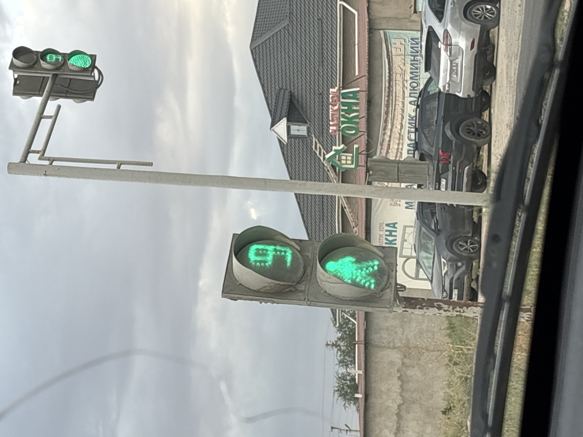 The image shows a traffic light with a countdown timer displaying "9" in green, and a green pedestrian signal. In the background, there are several cars parked, a building with a dark tiled roof, and a cloudy sky. A sign on the building reads "PLASTIK EDIL OKHA". The location is identified as Рахат ауылдық әкімдігі, Kazakhstan. There is also a faint, dark spiral shape visible in the sky, possibly a weather phenomenon or an artifact.