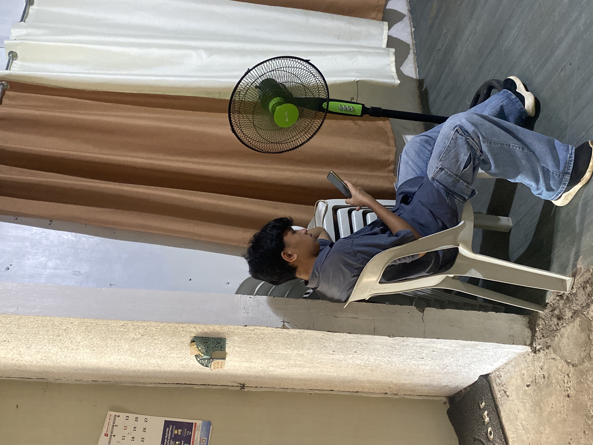 In Davao City, Philippines, a young man is seated on a white plastic chair, looking at his phone. He is wearing a blue collared shirt and light blue cargo pants. A black oscillating fan with a green accent is positioned behind him, and a stack of similar white chairs is to his left. The environment features a textured beige wall on the left, a concrete floor, and a doorway draped with brown and off-white curtains. A calendar and a small green organizer are mounted on the wall.