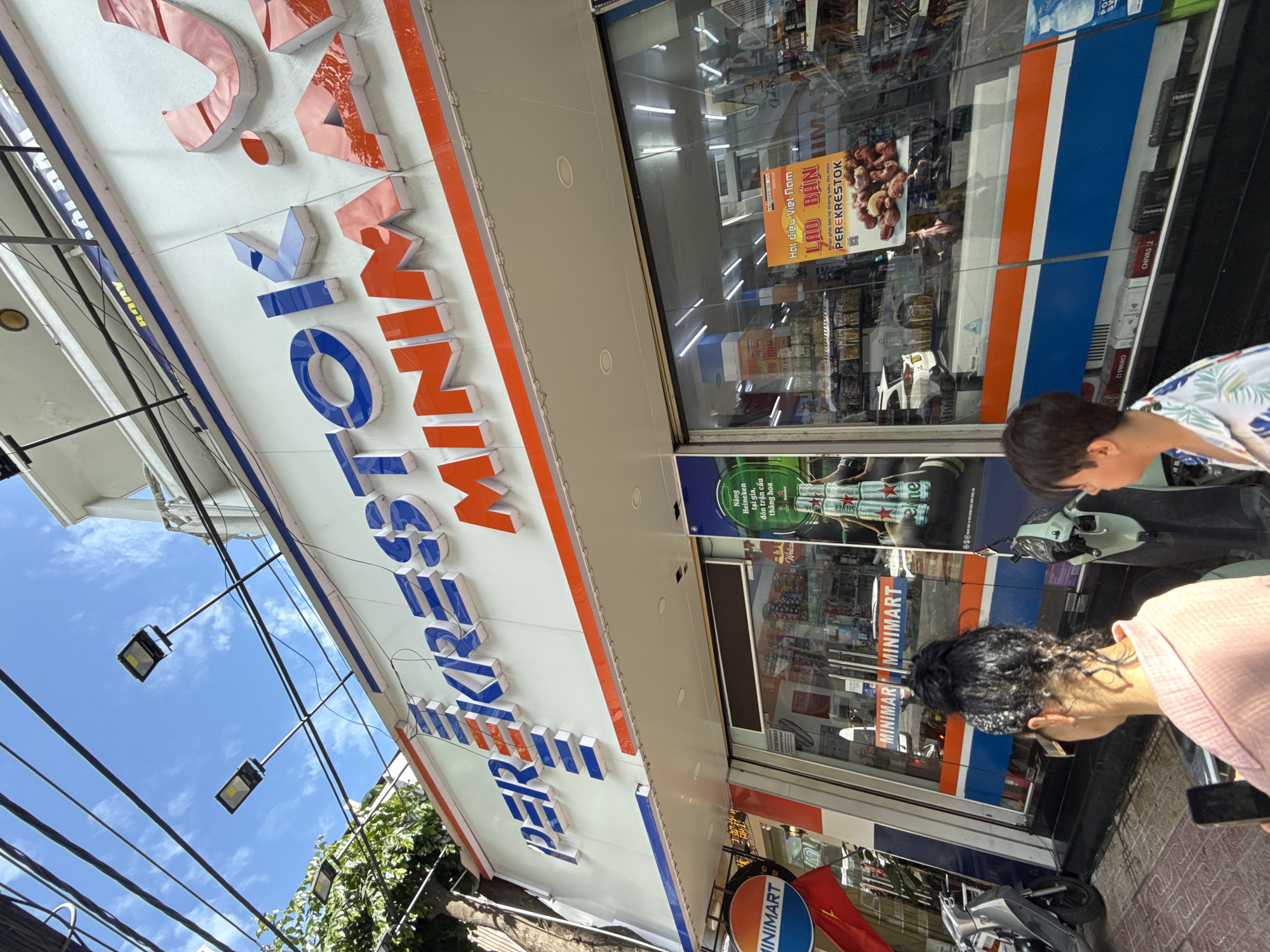 The image shows the exterior of a minimart store named "PER KRESTOK MINIMART" in Nha Trang, Vietnam. The store has a large white facade with the store name prominently displayed in blue and orange letters. The storefront features large glass windows, through which various products and shelves stocked with goods are visible. A promotional poster featuring nuts and Vietnamese text is displayed on the window. Outside the store, a person with dark curly hair tied up in a bun is seen from behind, wearing a pink top and holding a smartphone. Another person with dark hair is partially visible in the foreground. Several motorcycles are parked near the entrance. The street is paved with bricks, and overhead, a network of electrical wires crisscrosses against a blue sky with clouds. The overall scene depicts a typical street-level commercial establishment in a Vietnamese city.
