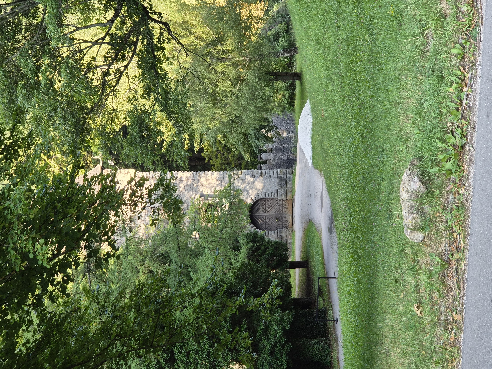 The image shows an exterior view of the Erzberg Castle in Eisenerz, Austria. The castle is made of stone and features a large wooden gate with intricate metalwork. It is surrounded by lush green trees and grass, with a paved path leading up to the entrance. The sunlight filters through the leaves, casting dappled shadows on the castle walls. There is a pile of white material on the grass to the right of the path, and a single rock in the foreground. The scene suggests a serene and historic setting.