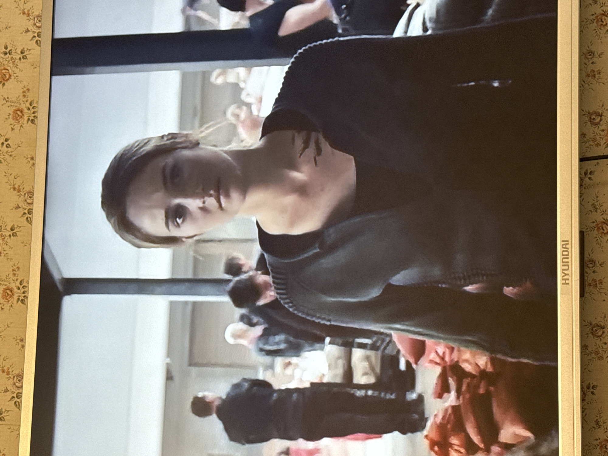 The image shows a scene from a film or television show being displayed on a Hyundai monitor. A fair-skinned woman is the central figure, seen from the chest up. She has light brown hair and is wearing a black top or jacket. There appears to be a dark tattoo or marking on her upper chest or shoulder. In the background, a group of people is visible, some in dark clothing and some in lighter colors, possibly costumes. The scene seems to be set in a large, possibly public, space. The monitor is positioned against a floral wallpaper. The image does not provide any information on the specific location in Mytishchi, Russia, nor does it show any recognizable landmarks or characteristics that would allow for determination of location in the city.