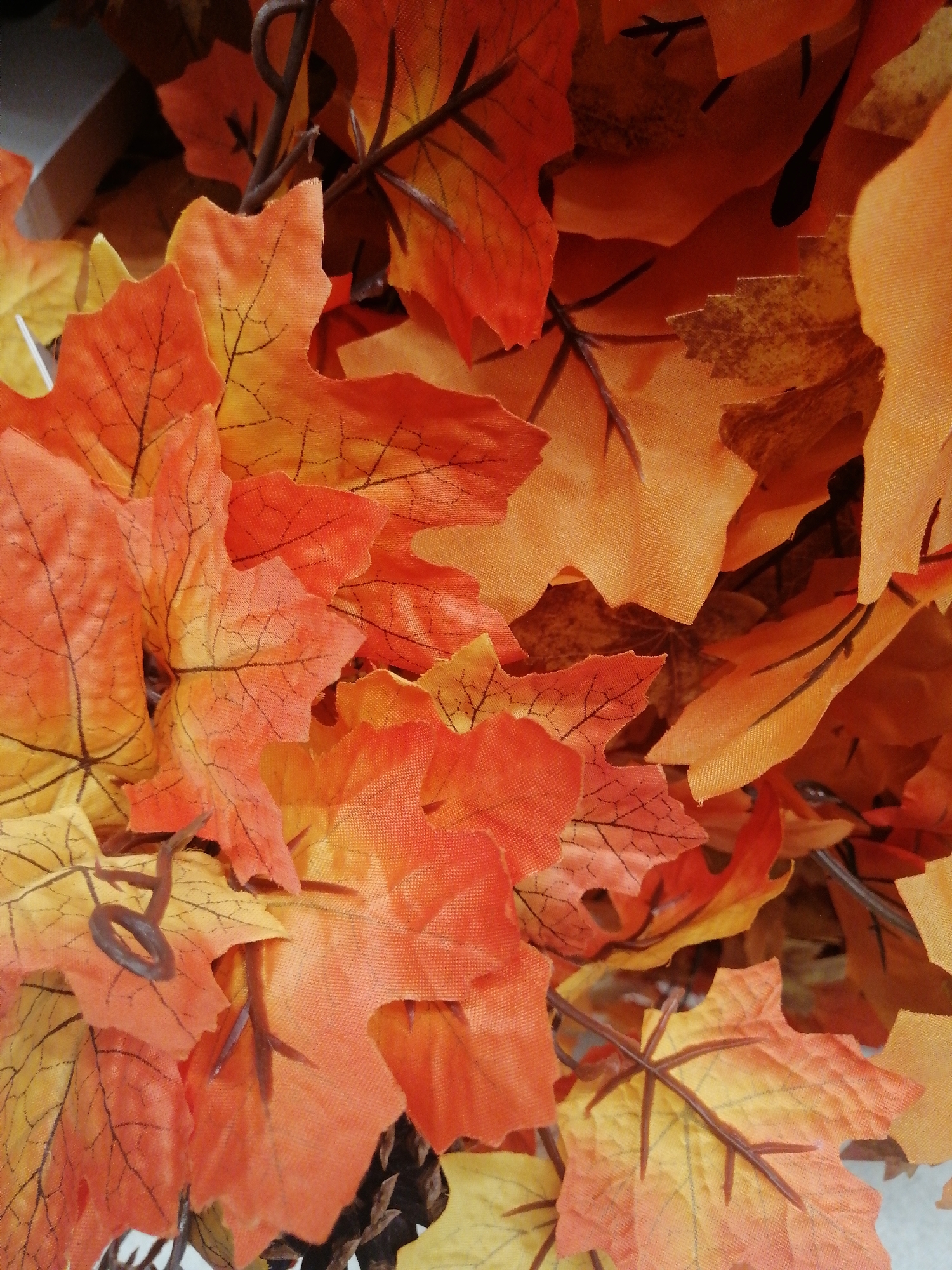 The image displays a close-up view of a collection of artificial autumn leaves, predominantly in shades of orange, red, and yellow. The leaves have a fabric texture with printed vein details. Some brown, wire-like stems are visible, connecting and supporting the leaves, suggesting they are part of a garland or decorative arrangement. The lighting is somewhat harsh, creating bright highlights and deep shadows, indicating an artificial light source or direct sunlight. There are no people or animals present in the image. The background is mostly obscured by the leaves, but appears to be a darker, indistinct area. This scene does not contain any identifiable landmarks or elements that would allow for its specific location to be determined within the city of Kremenchuk, Ukraine.