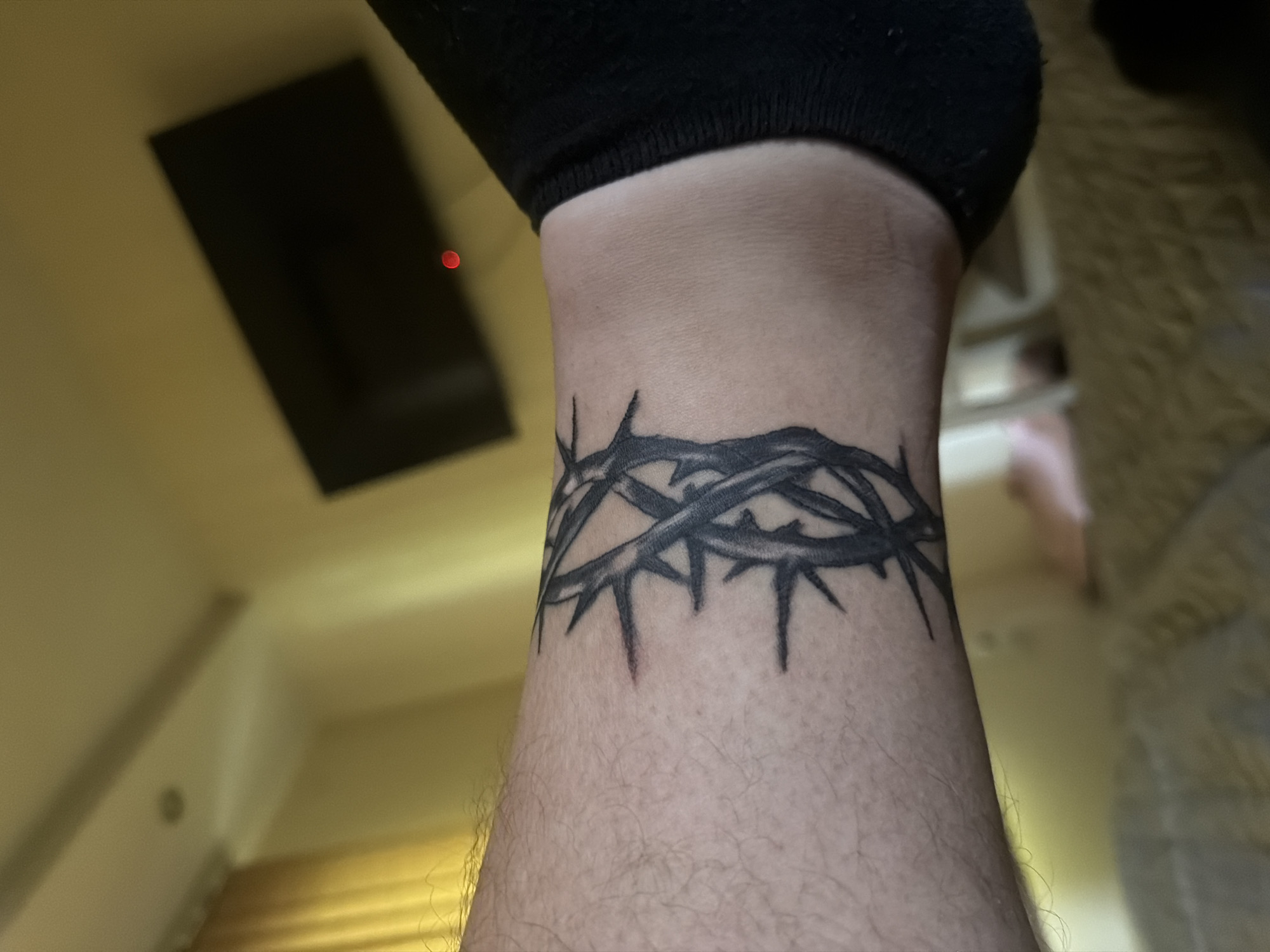 A close-up shot captures a person's lower leg or arm, most likely an ankle, adorned with a striking black tattoo. The tattoo depicts a crown of thorns, intricately detailed and wrapping around the limb. Above the tattoo, the edge of a black sock or sleeve is visible. The skin is fair-toned and covered with fine body hair.
The background is out of focus, indicating an indoor setting under artificial lighting, which casts a warm, soft glow across the scene. In the upper left corner, a dark, rectangular object, possibly a television or a framed item, reflects a small, distinct red light. Other blurred elements in the background suggest a wall or ceiling above and an indistinct patterned surface, possibly a rug or floor, to the right. No specific activity is depicted, and the image appears to be a static capture focused on showcasing the tattoo. There is no visible text in the image. The setting is consistent with a home interior in Shumen, Bulgaria.