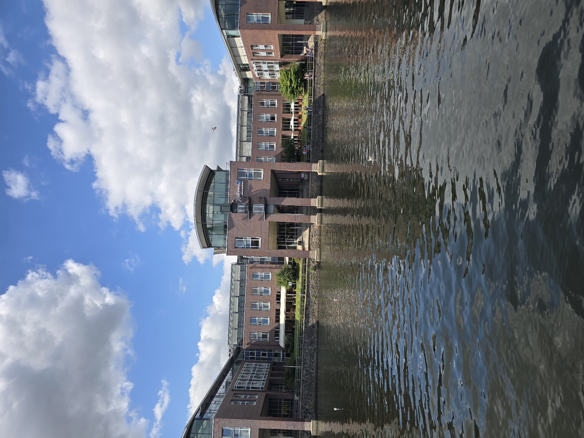 This is a medium-shot, eye-level, outdoor, vertical photograph taken from the water, looking towards the Radisson Blu Senator Hotel in Lübeck, Germany. The building, constructed of red brick and featuring large glass windows and a curved glass roof, faces a canal. Several people are visible walking along the path by the canal in front of the hotel. A few seagulls are present in the water. The sky is blue with scattered white clouds. The water of the canal reflects the sky and the building. The scene captures the hotel on a sunny day with moderate cloud cover.