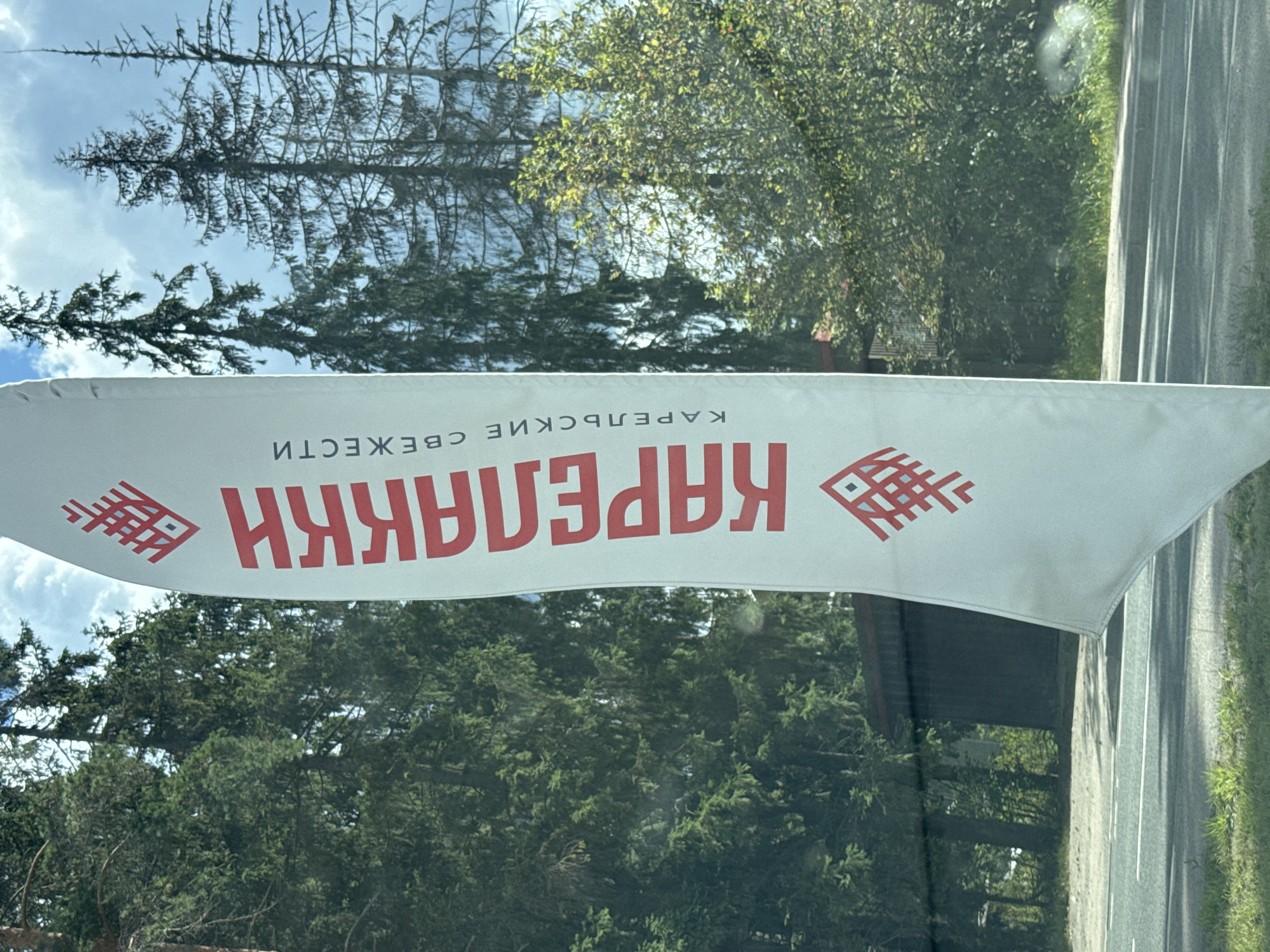 The image shows a white banner with red lettering and symbols, likely in Yakkima, Russia. The banner is vertical and features a decorative pattern at the top and bottom, and the word "КАРЕЛАККИ" in large red letters down the center. Below this, in smaller red letters, is the word "СВЕЖЕСТИ". The banner is positioned outdoors, with a backdrop of tall, green coniferous trees and a bright blue sky with scattered clouds. In the foreground, a road with white lines is visible, bordered by grass and a tree line. A dark, possibly wooden structure is partially obscured by trees behind the banner. The scene suggests a commercial or promotional display.