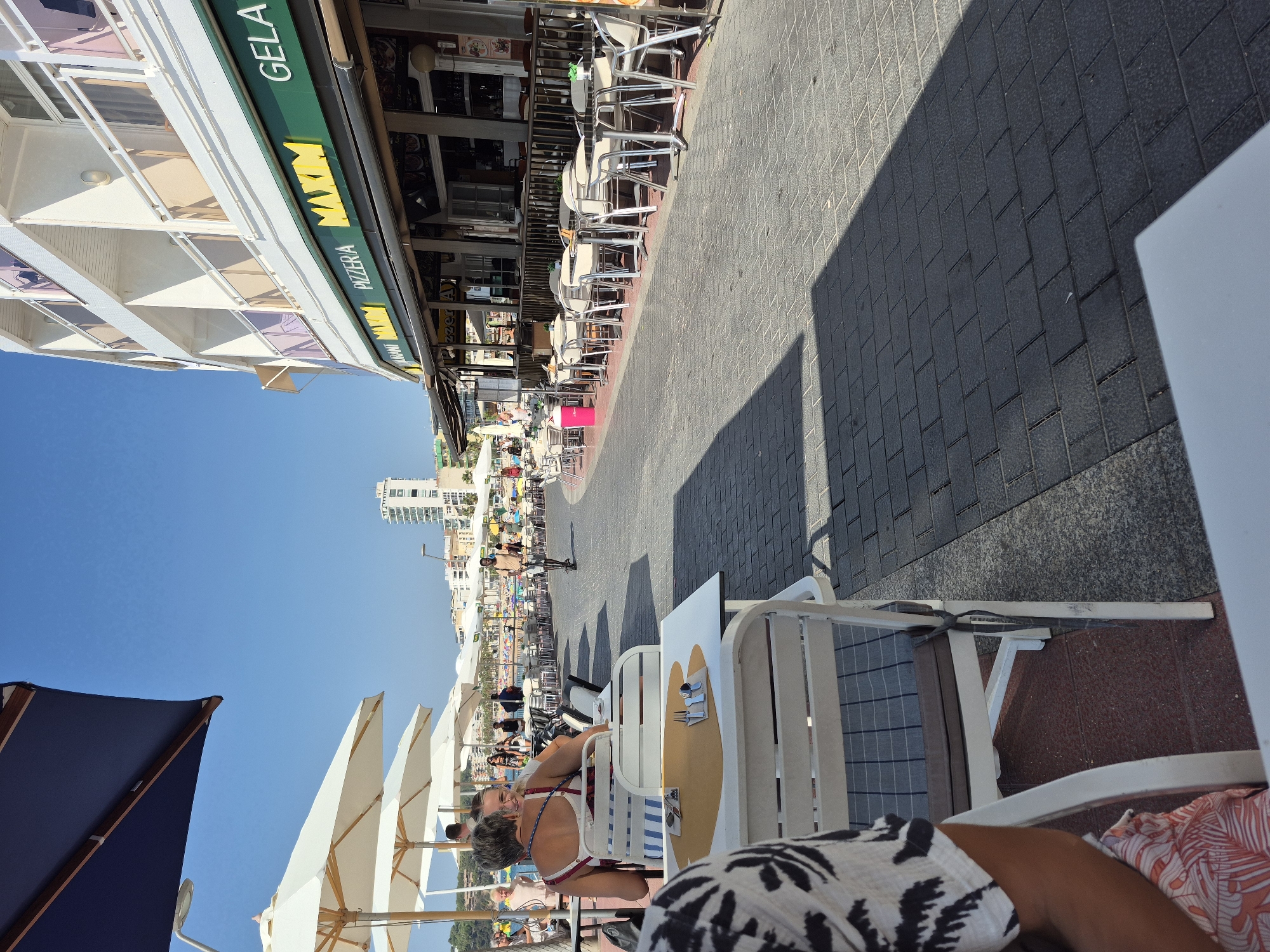 This is an outdoor photograph taken on a sunny day in Lloret De Mar, Spain, likely along the promenade by the beach. In the foreground, a white table and chairs are set up, with a partially visible person seated in a chair on the left, wearing a patterned shirt and a pink and white floral skirt. Another person is visible in the mid-ground, seated at a table with a blue and white striped cushion. Beyond the seating area, a pedestrian walkway paved with grey and dark grey brick-like tiles stretches into the distance. Along the walkway, numerous outdoor tables and chairs belonging to restaurants are arranged. A building with multiple balconies and white railings runs along the right side of the promenade, with a prominent green awning above the ground floor businesses. A sign with yellow lettering reads "MAXIM PIZZERIA GELATO". In the background, more beach umbrellas and people are visible, along with buildings and a tall white building further inland. A person is seen riding an electric scooter on the walkway in the mid-ground. The sky is clear and blue.